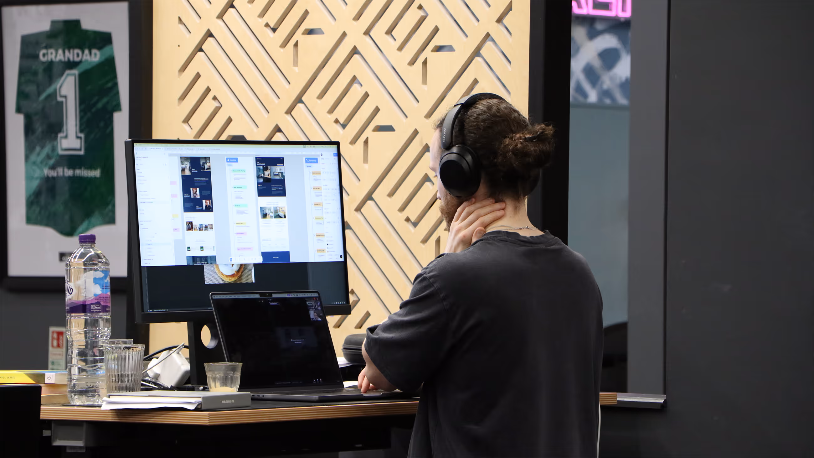 Person with headphones working on laptop and desktop showing website design on desk with water bottle.