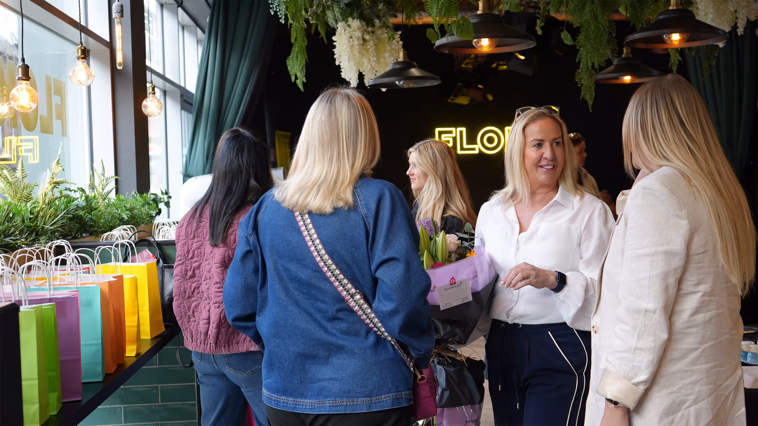 Women chatting inside a flower shop with colorful gift bags and floral arrangements visible.