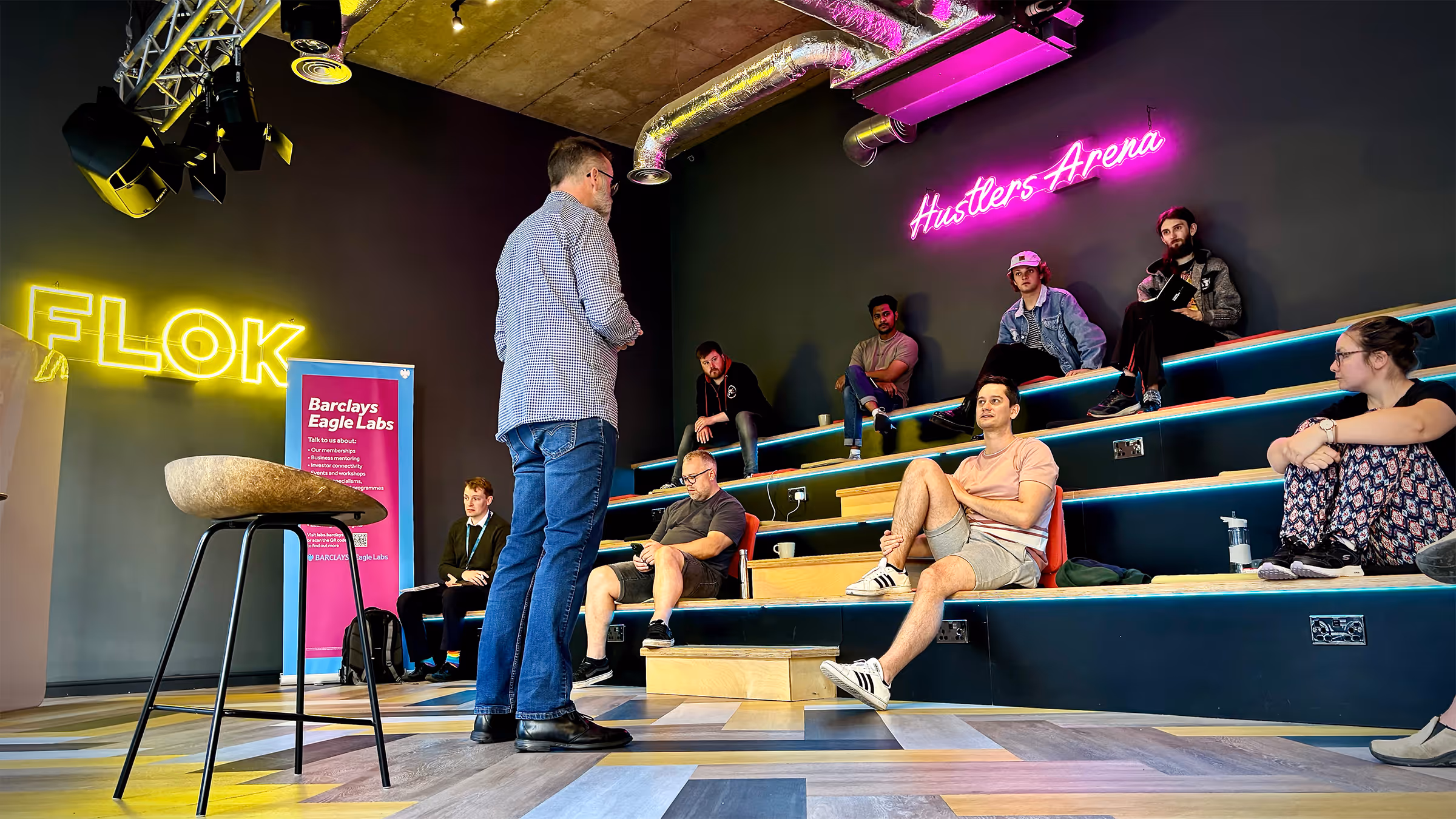 Man standing and talking to seated group on tiered benches under neon signs reading FLOK and Hustlers Arena.