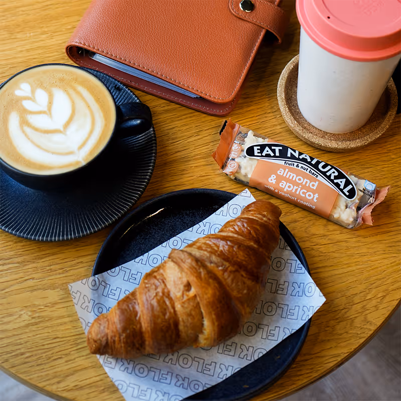 Croissant on a black plate with latte art coffee, almond apricot bar, red notebook, and pink-lid cup on wood table.