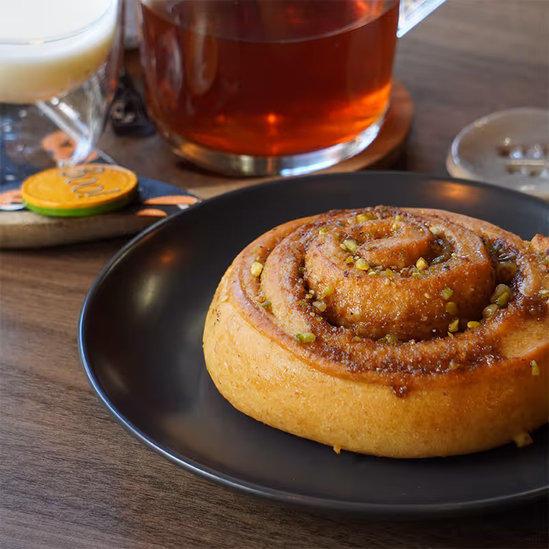 Cinnamon roll topped with chopped nuts on a black plate beside a glass cup of tea.