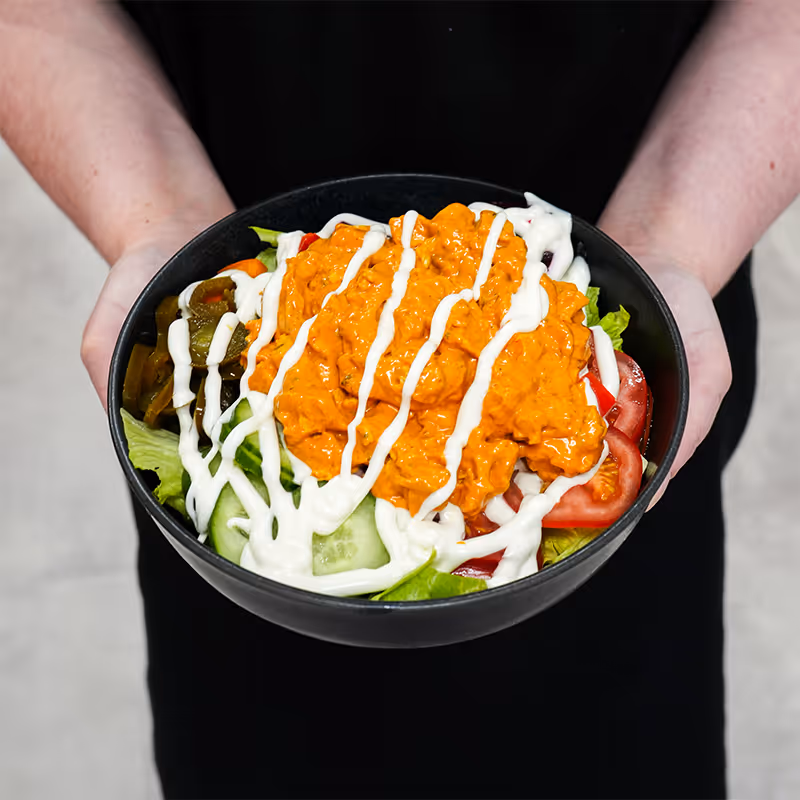 Person holding bowl of salad with lettuce, tomato, cucumber, pickles, orange sauce, and drizzled white dressing.