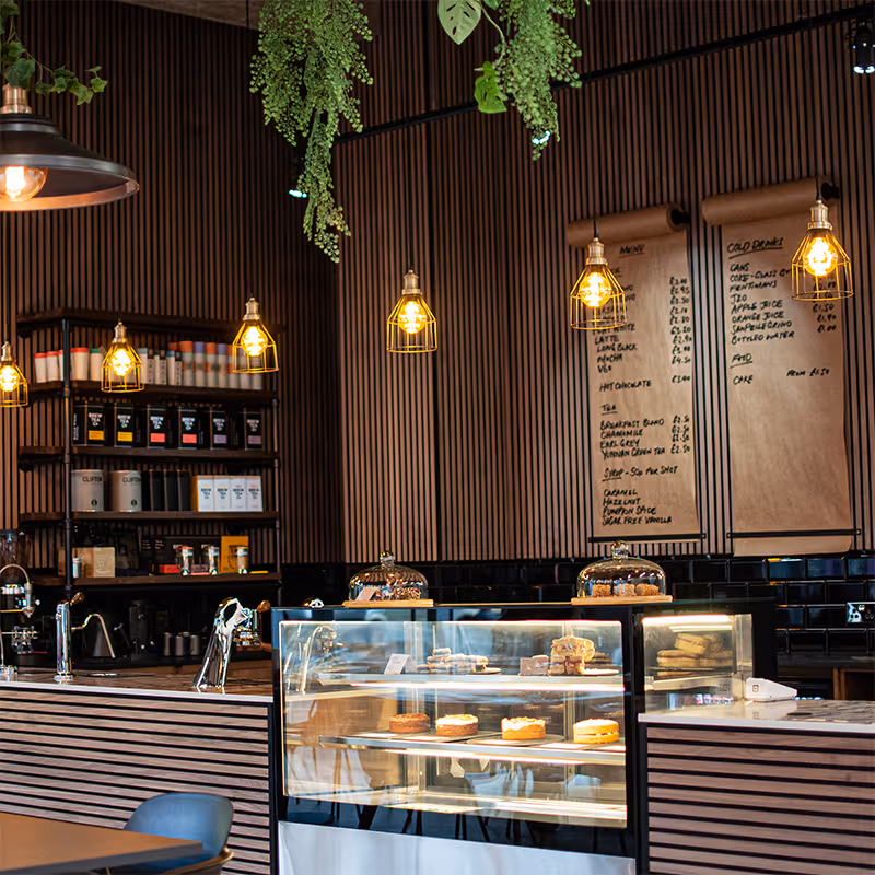 Interior of a modern cafe with hanging lights, plants, a display case of cakes, and a handwritten menu on the wall.