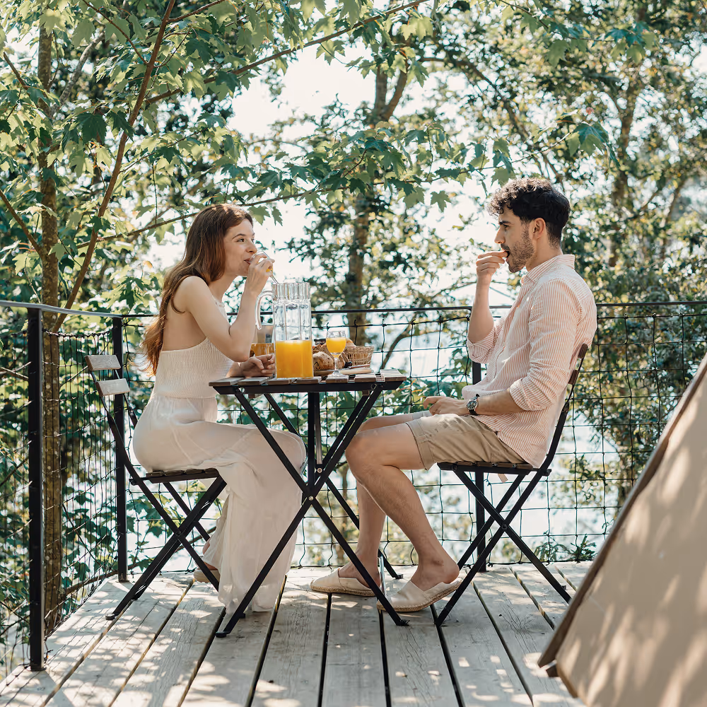 Couple sits at a cafe table eating lunch, surrounded by trees on all sides.