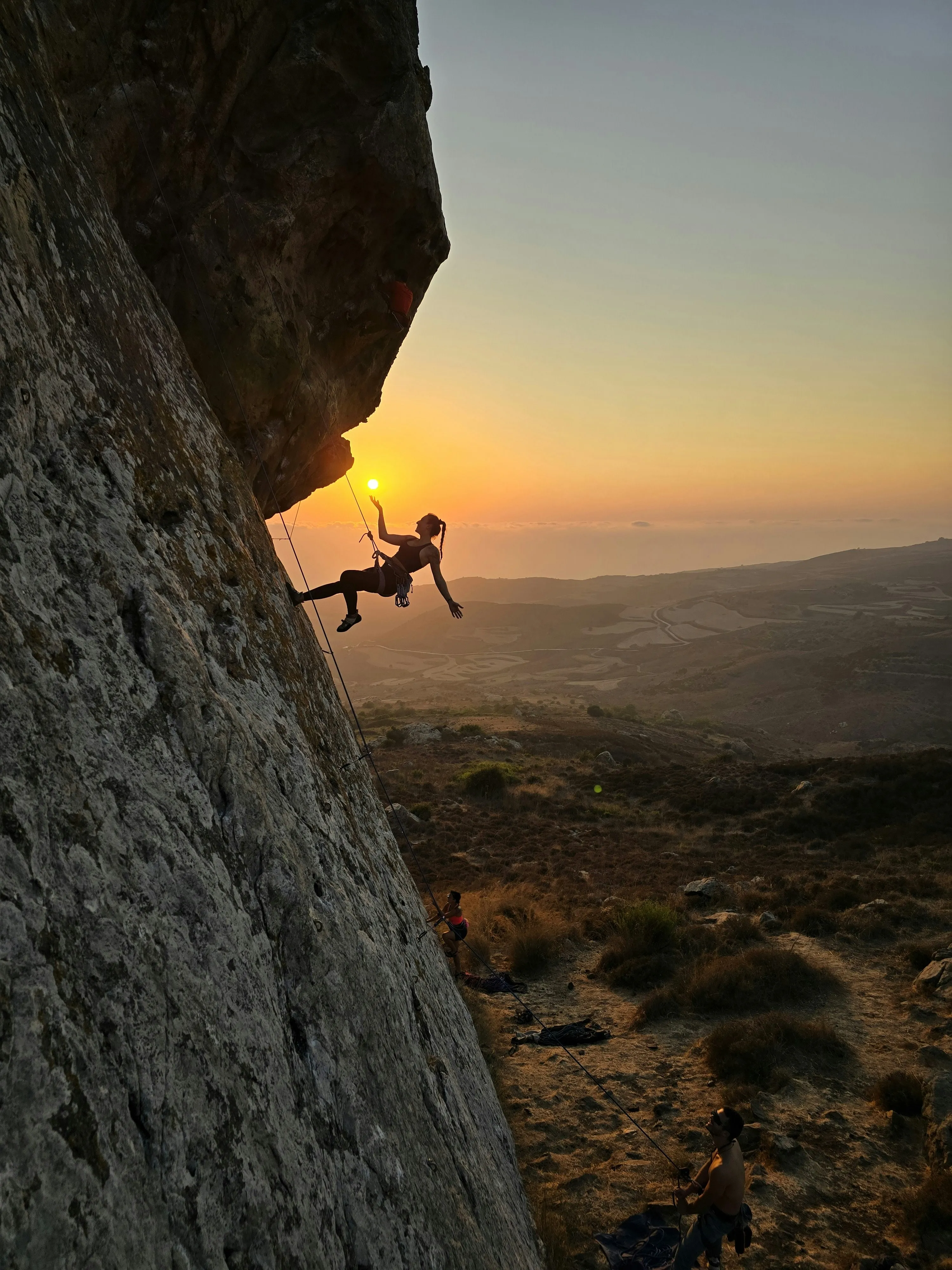 A woman climbs up a boulder with the rising sun.