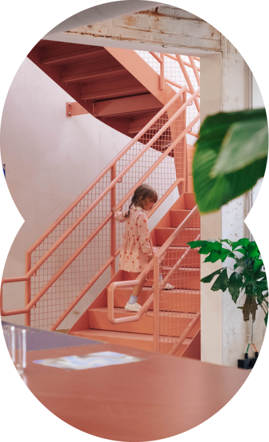 Young girl wearing a pink dress climbing a peach-colored metal staircase indoors.