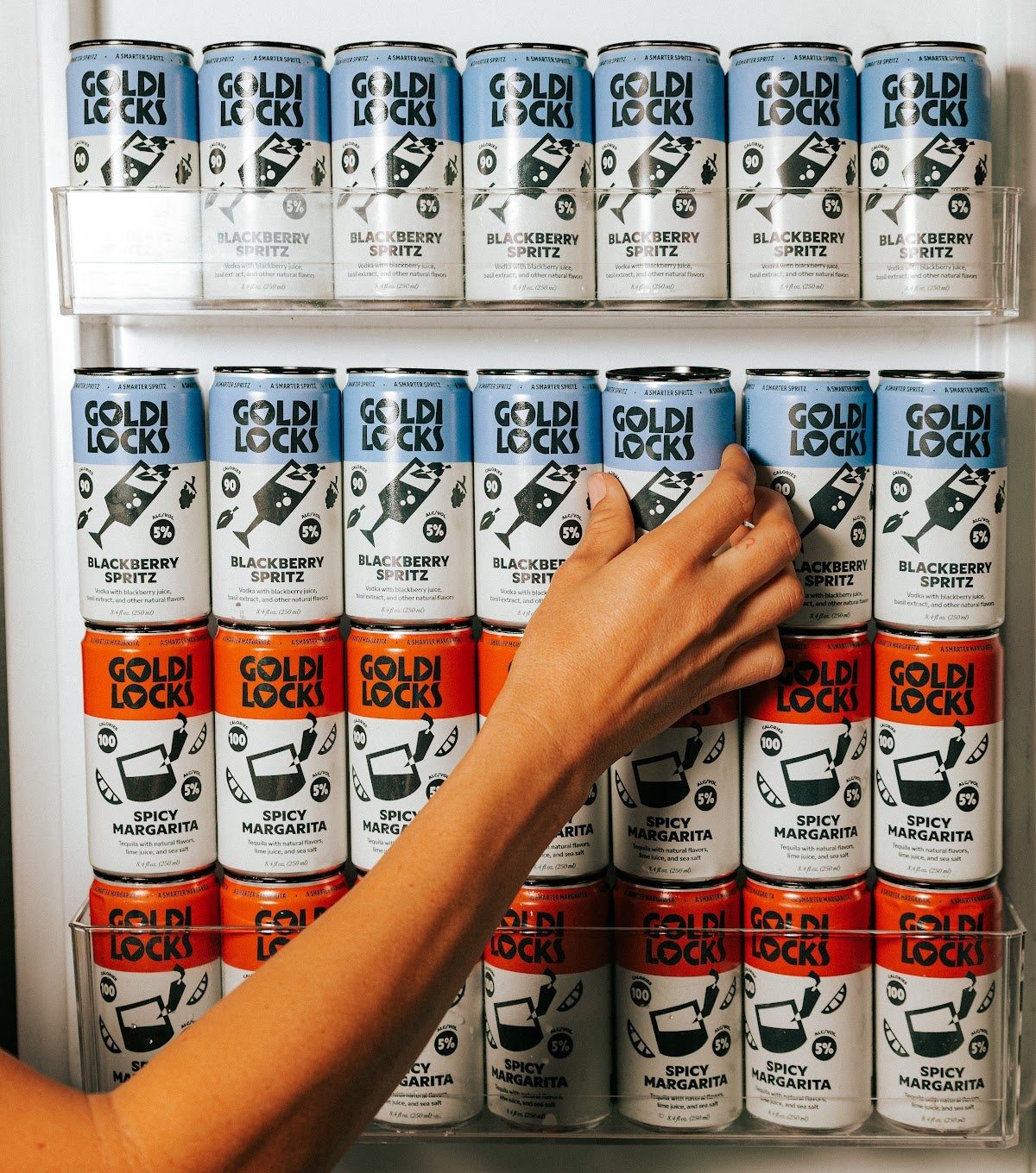 A hand grabbing a Blackberry Spritz from a fridge stocked with Spicy Margaritas and Blackberry Spritz