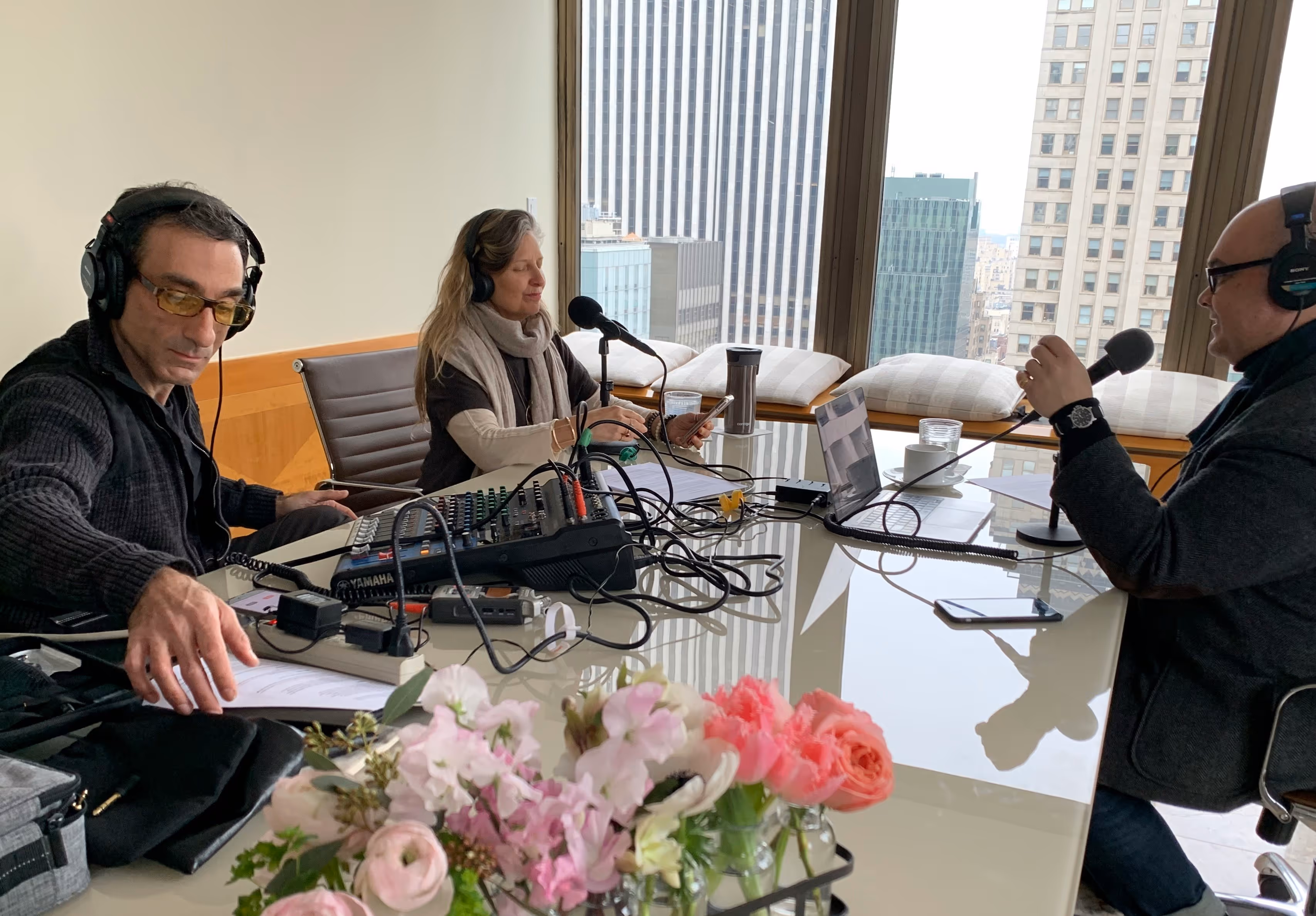 Three people with headphones and microphones recording a podcast in a room with large city-view windows.