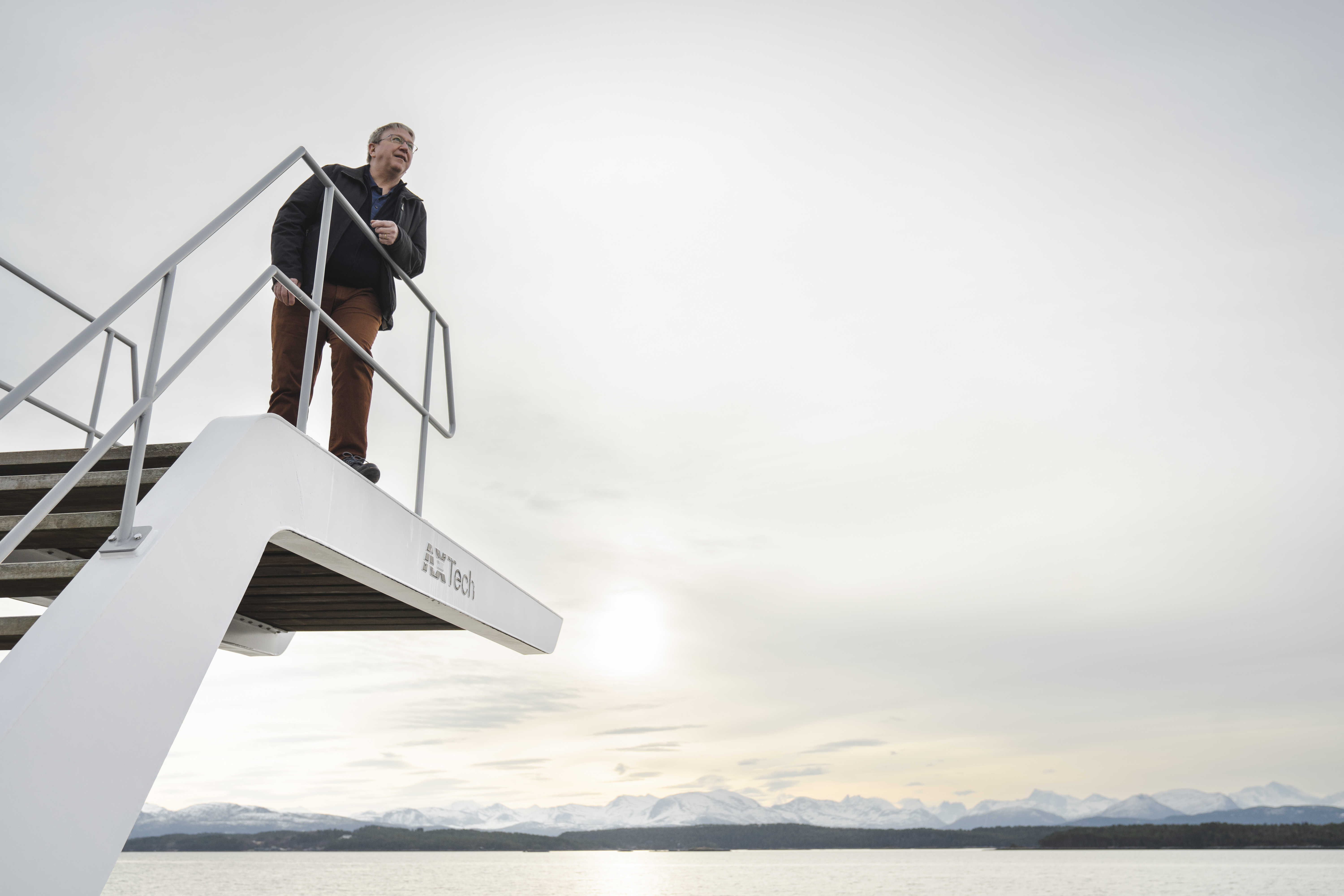 Man standing on a diving platform overlooking a calm body of water with snow-capped mountains in the distance under a cloudy sky.