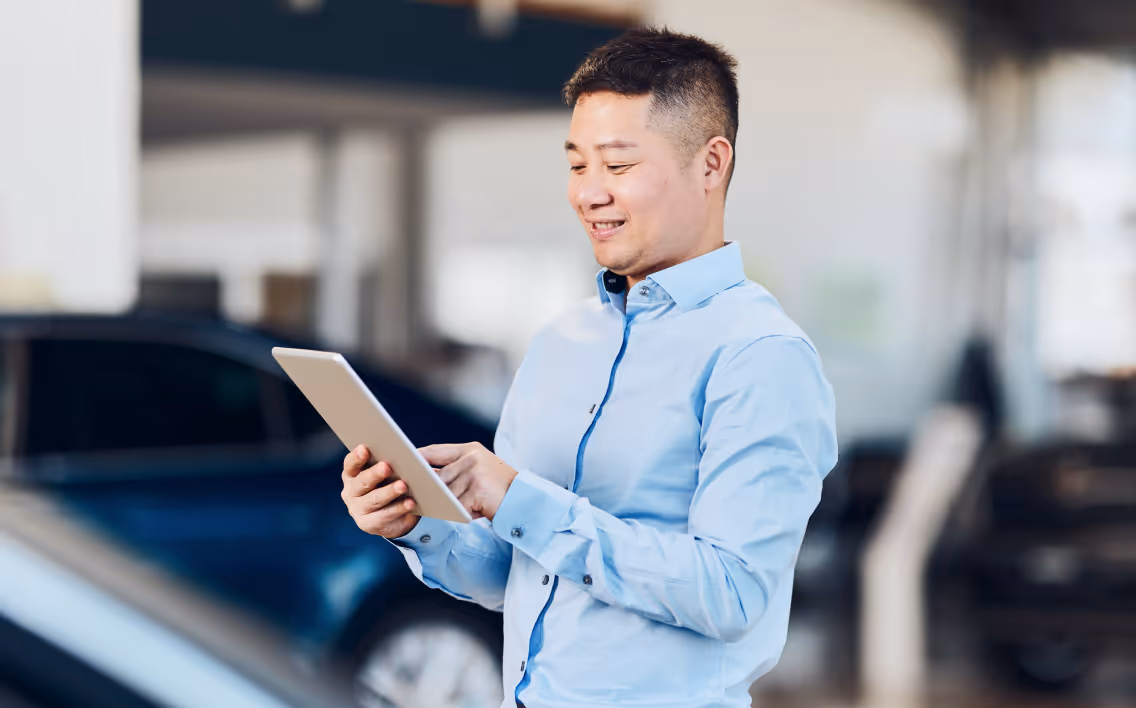 asian man in a car dealership looking down and smiling at an ipad