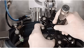 Gloved hands adjusting components on a laboratory or industrial machine with various pipes and metal parts.