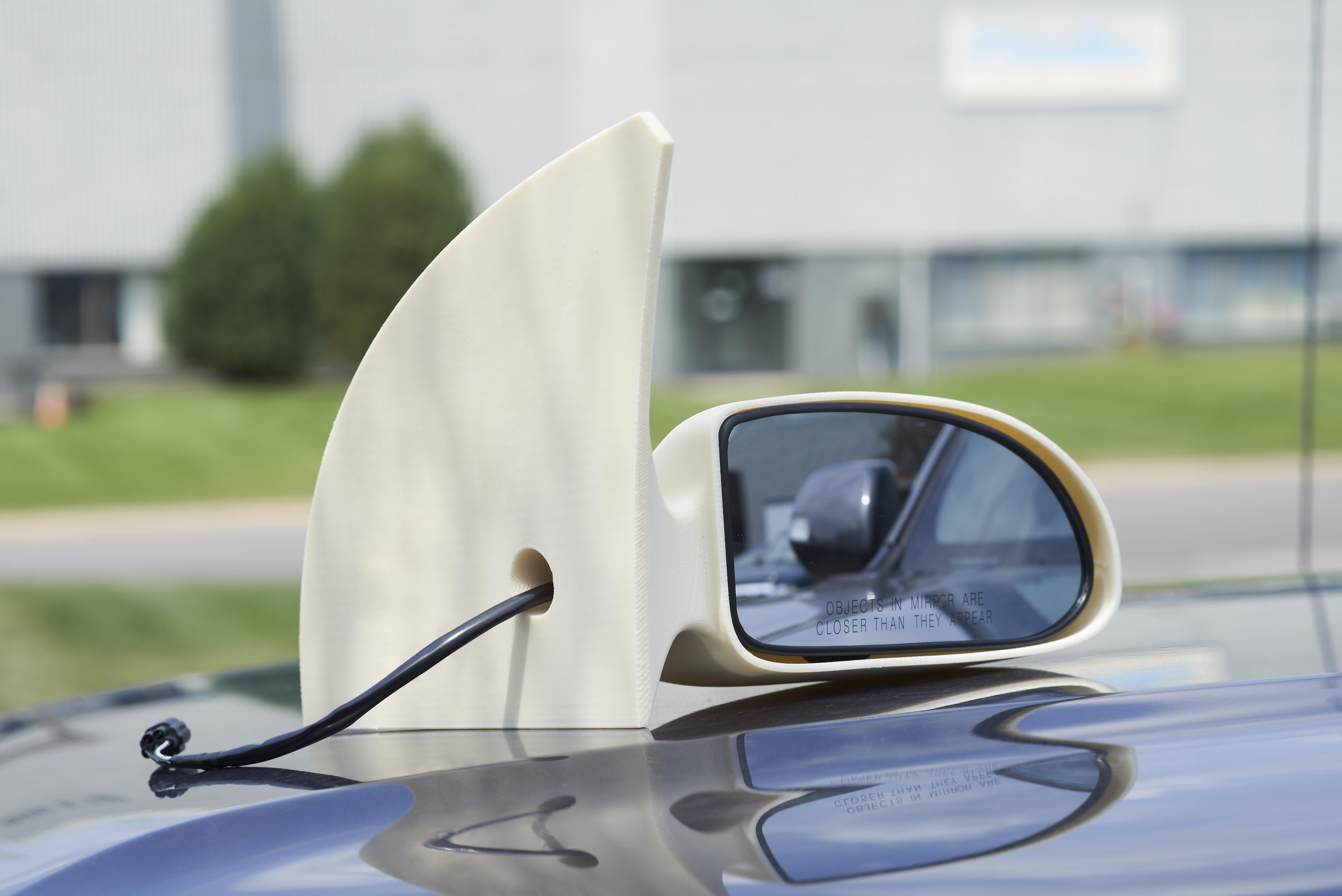 White car side mirror mounted on a blue car hood, reflecting a blurred vehicle side and displaying the warning text 'Objects in mirror are closer than they appear.'