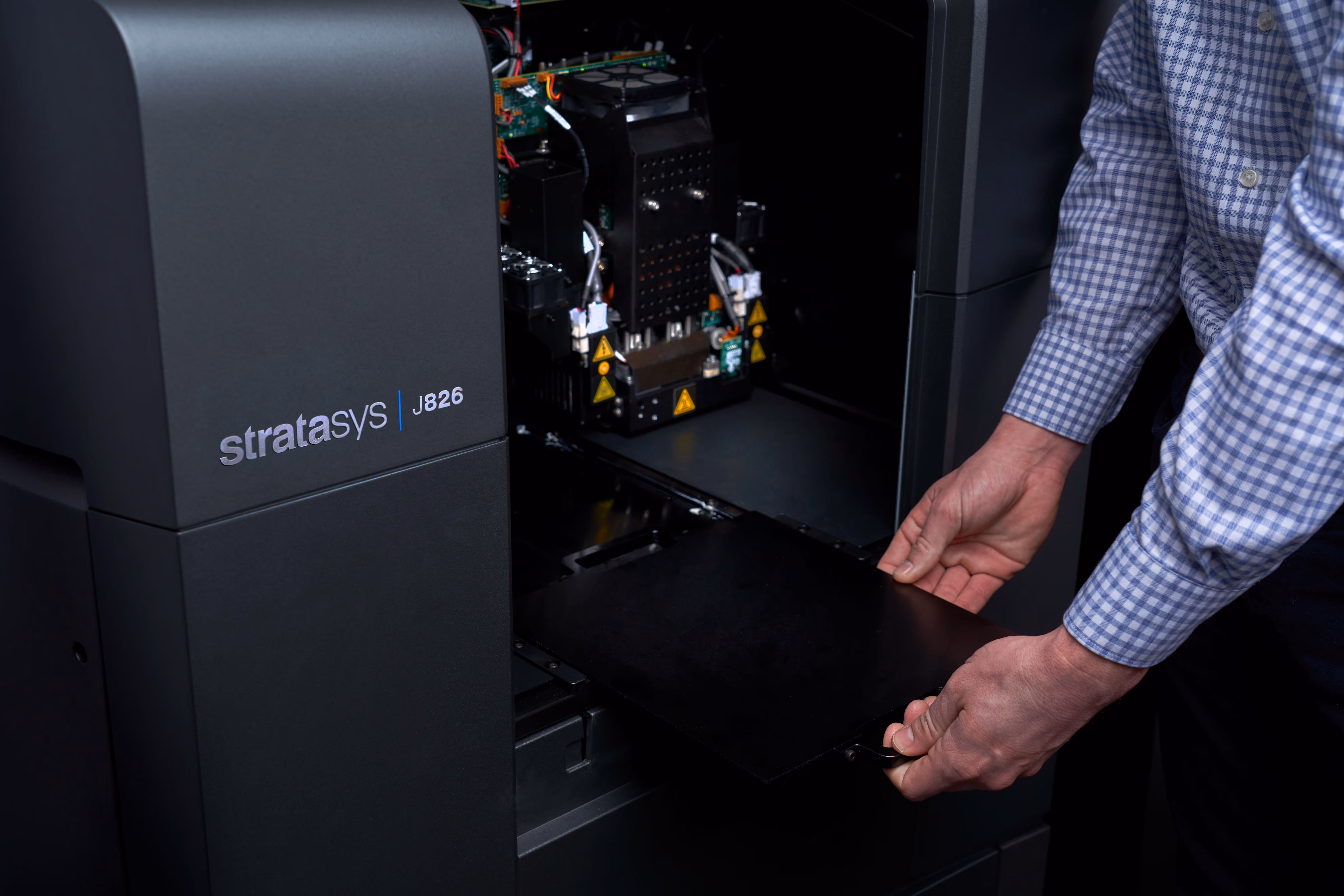 Person removing a black print tray from an open Stratasys J826 3D printer.