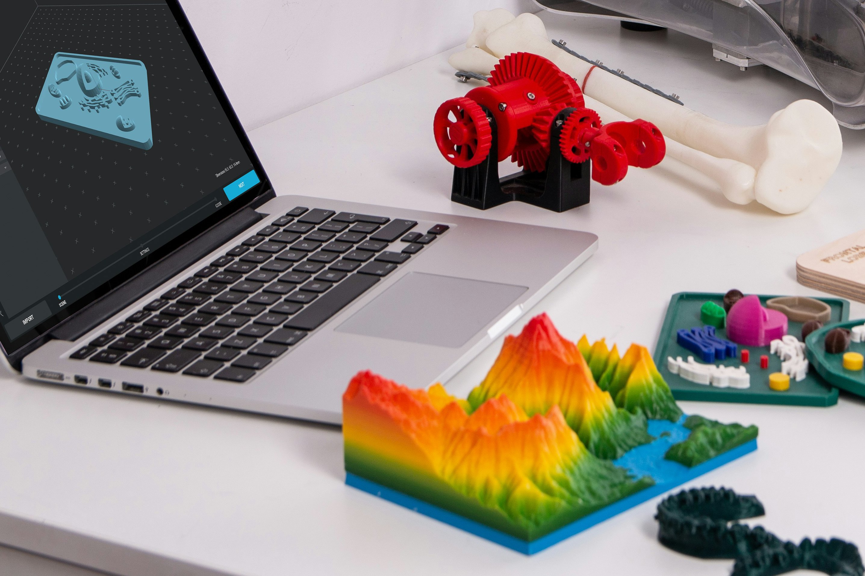Desk with a laptop showing 3D design software and various colorful 3D printed models including a topographic map, red gears, and anatomical bones.