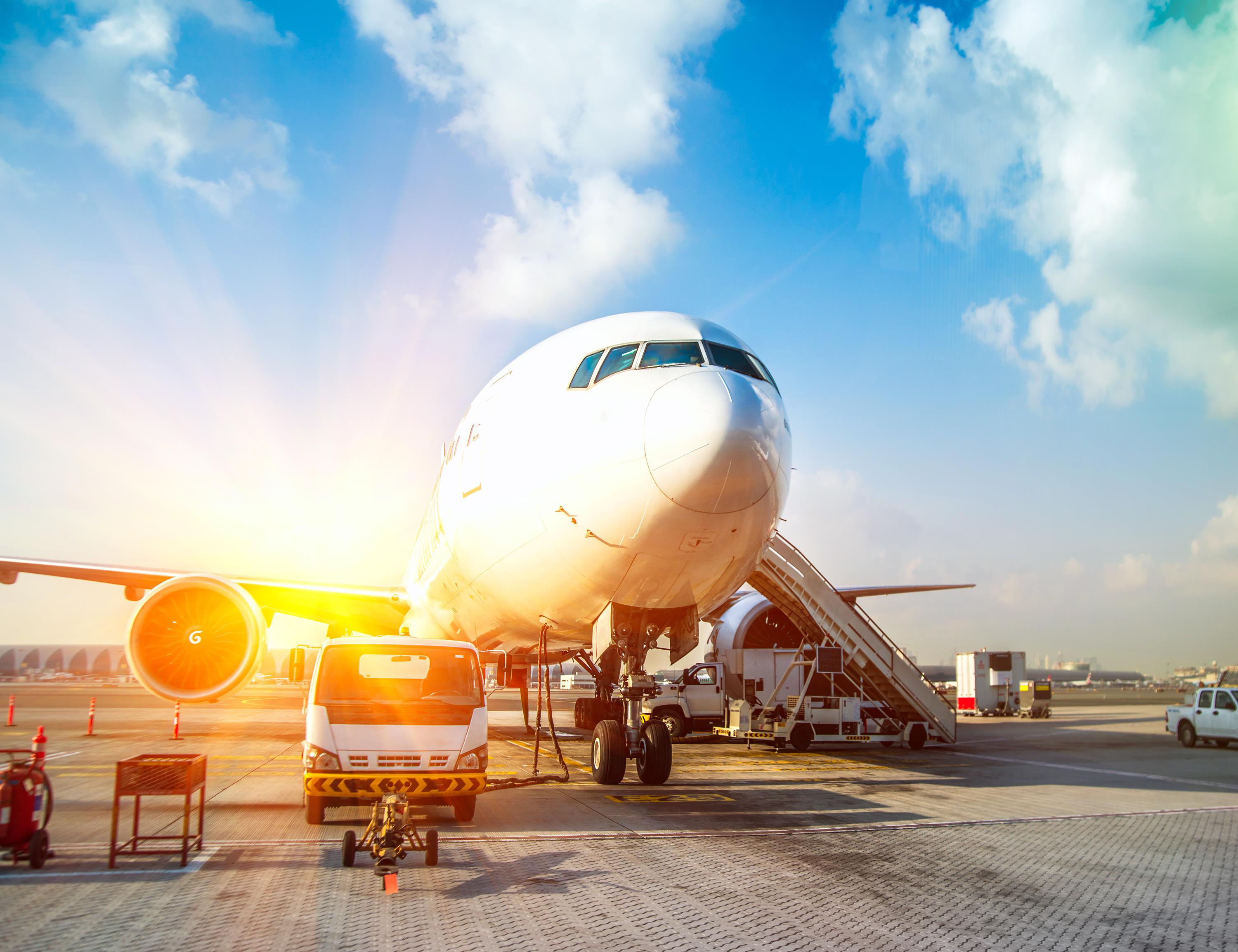Commercial airplane parked at an airport gate with ground service vehicle and boarding stairs under a sunny sky.