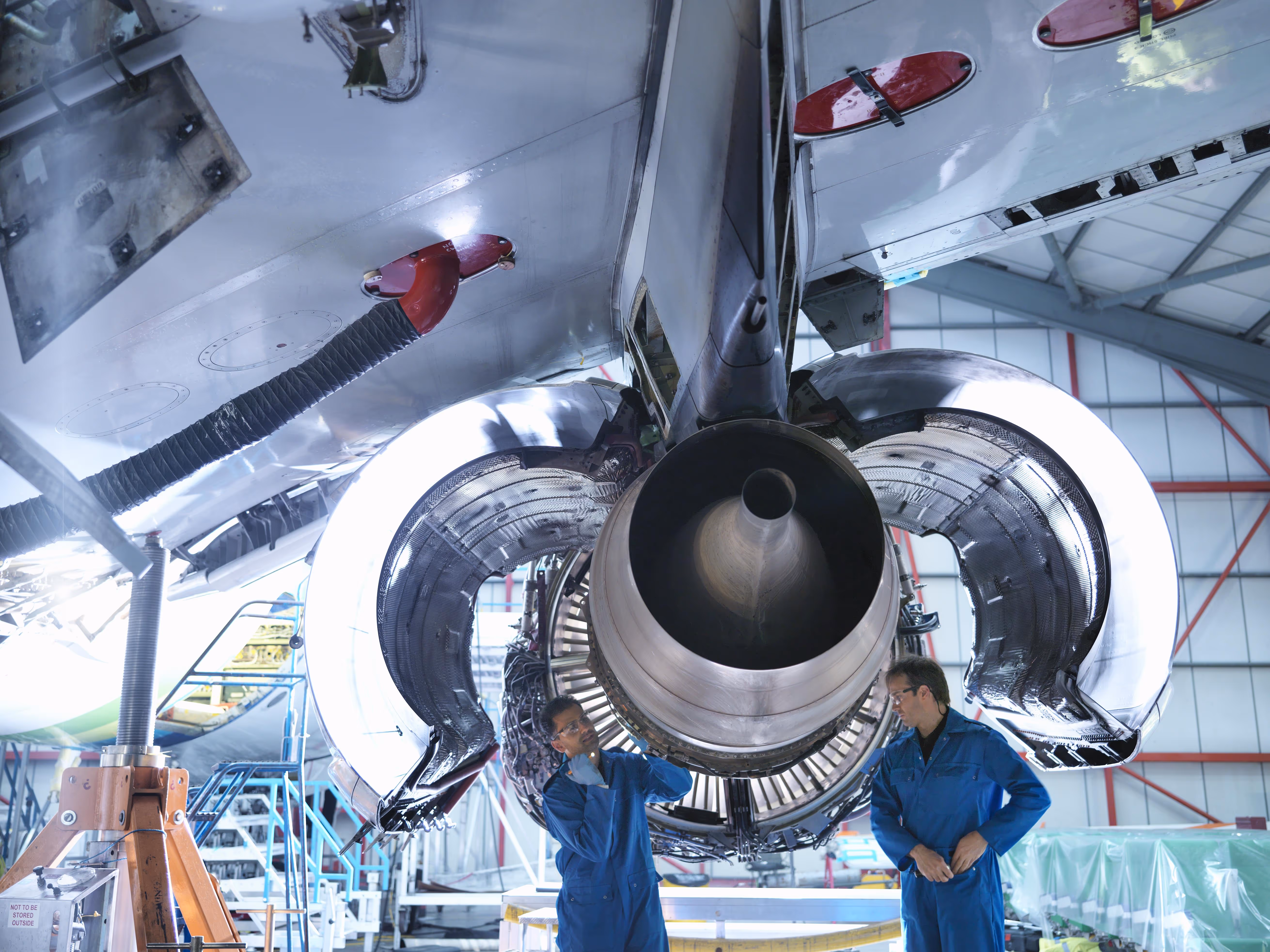 Two aircraft technicians in blue coveralls inspecting a large jet engine inside an aircraft maintenance hangar.