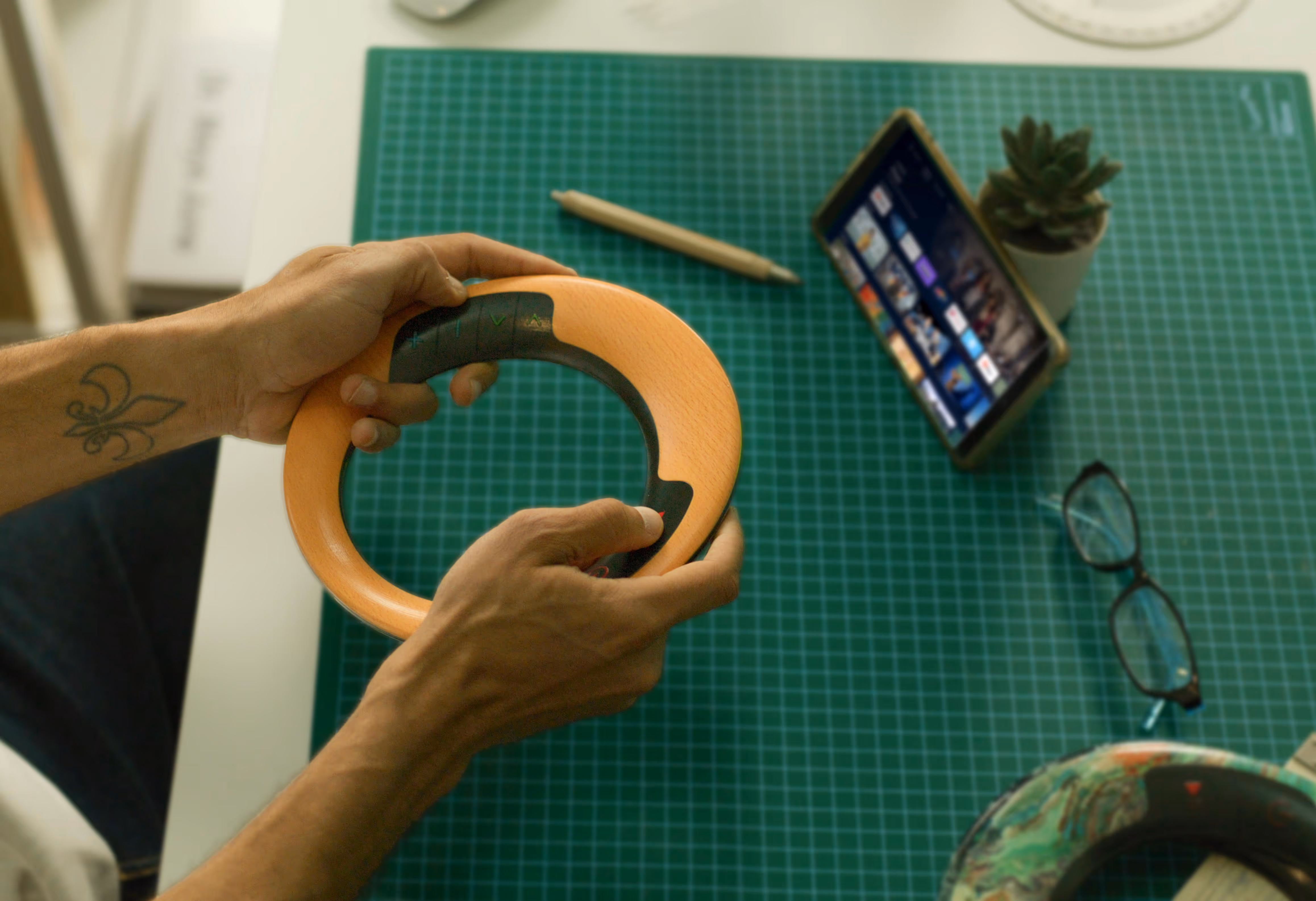 Person holding a wooden steering wheel-shaped remote control over a green cutting mat with a tablet, pen, glasses, and small potted plant nearby.