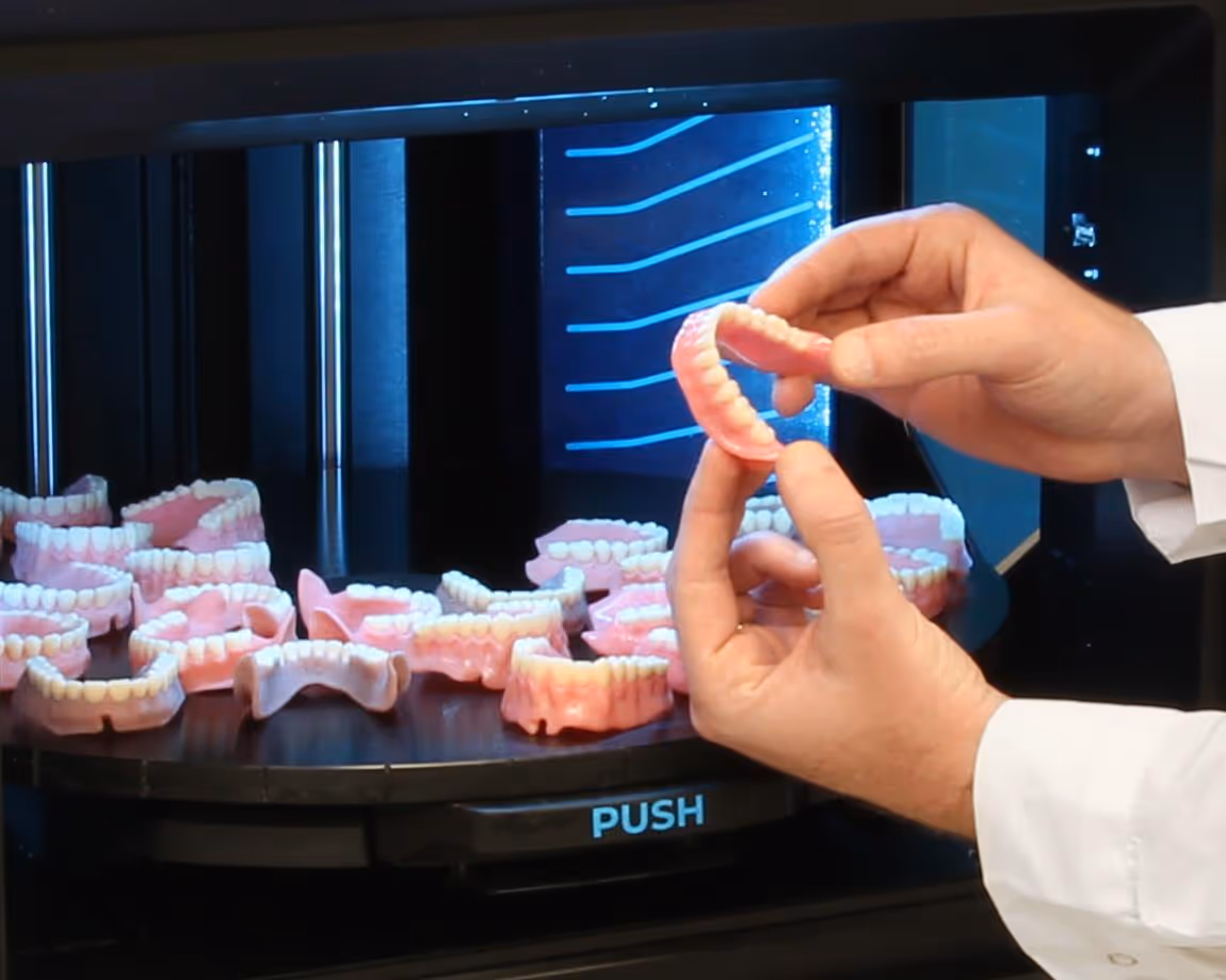 Hands holding a dental prosthetic with multiple dental models arranged on a rotating platform inside a 3D printer.