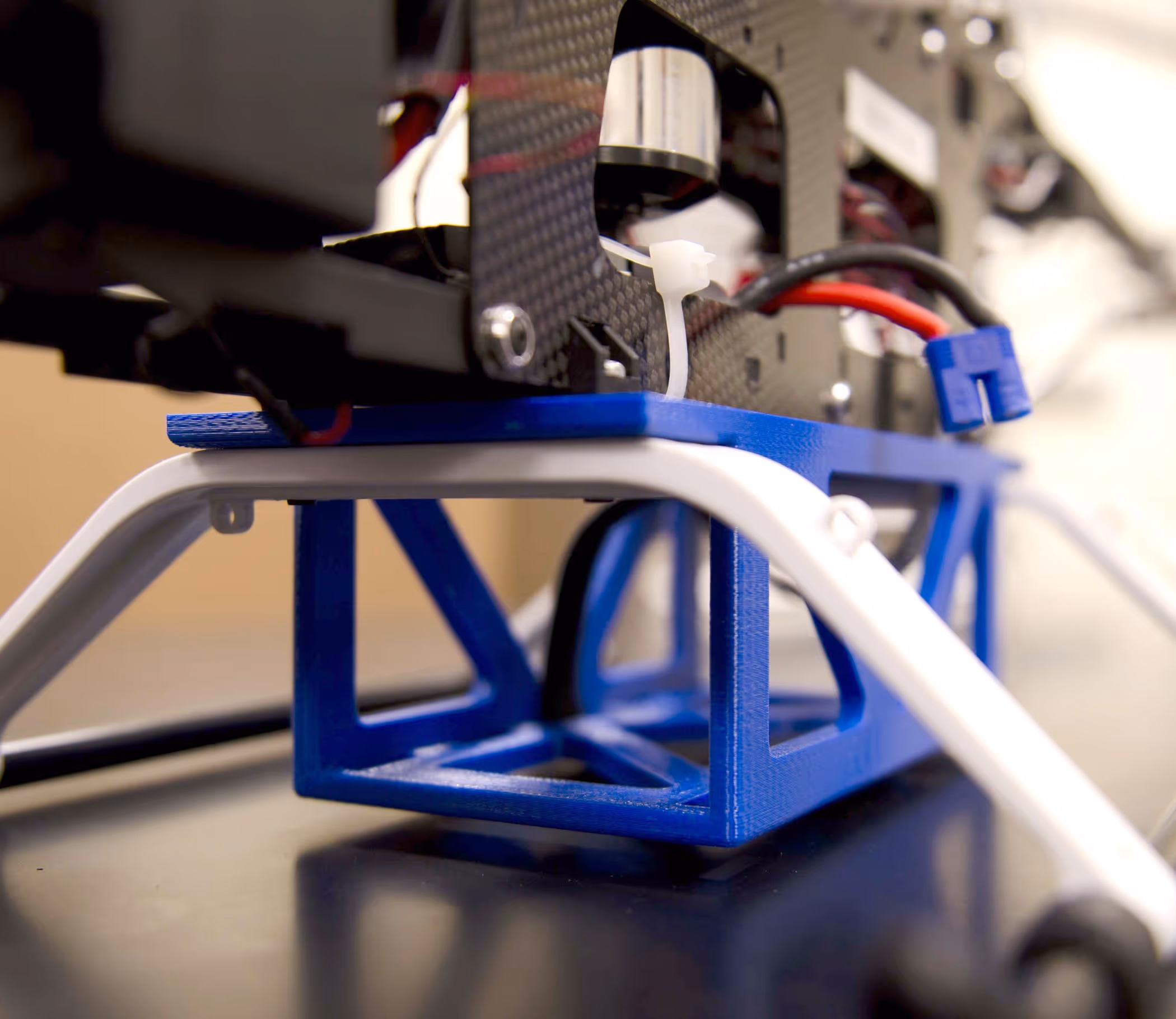 Close-up of a drone landing gear with white legs and a blue 3D-printed mount under a carbon fiber frame with cables.