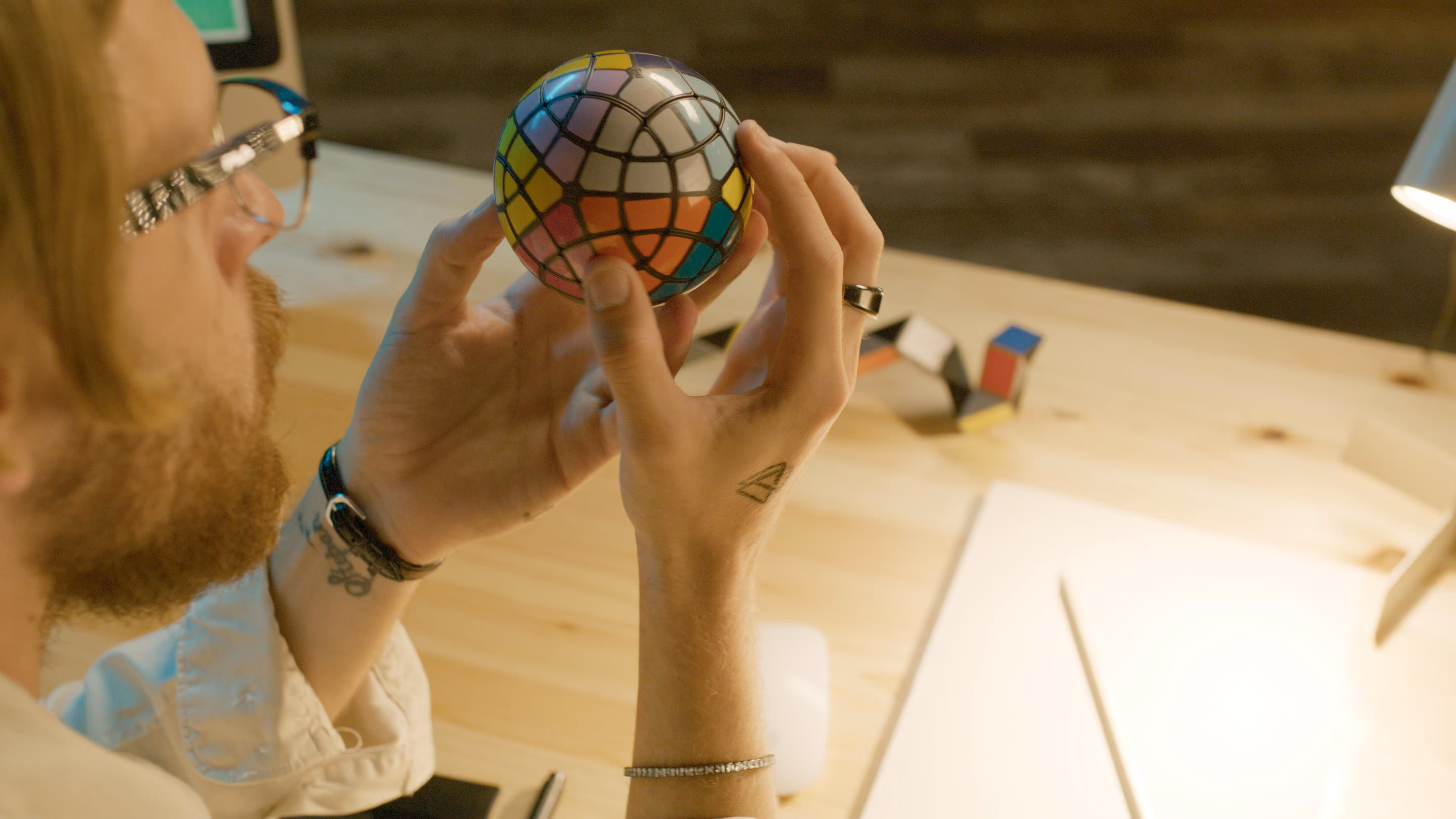 Man with glasses and tattoos holding a multicolored complex twisty puzzle ball at a wooden desk with papers and a lamp.