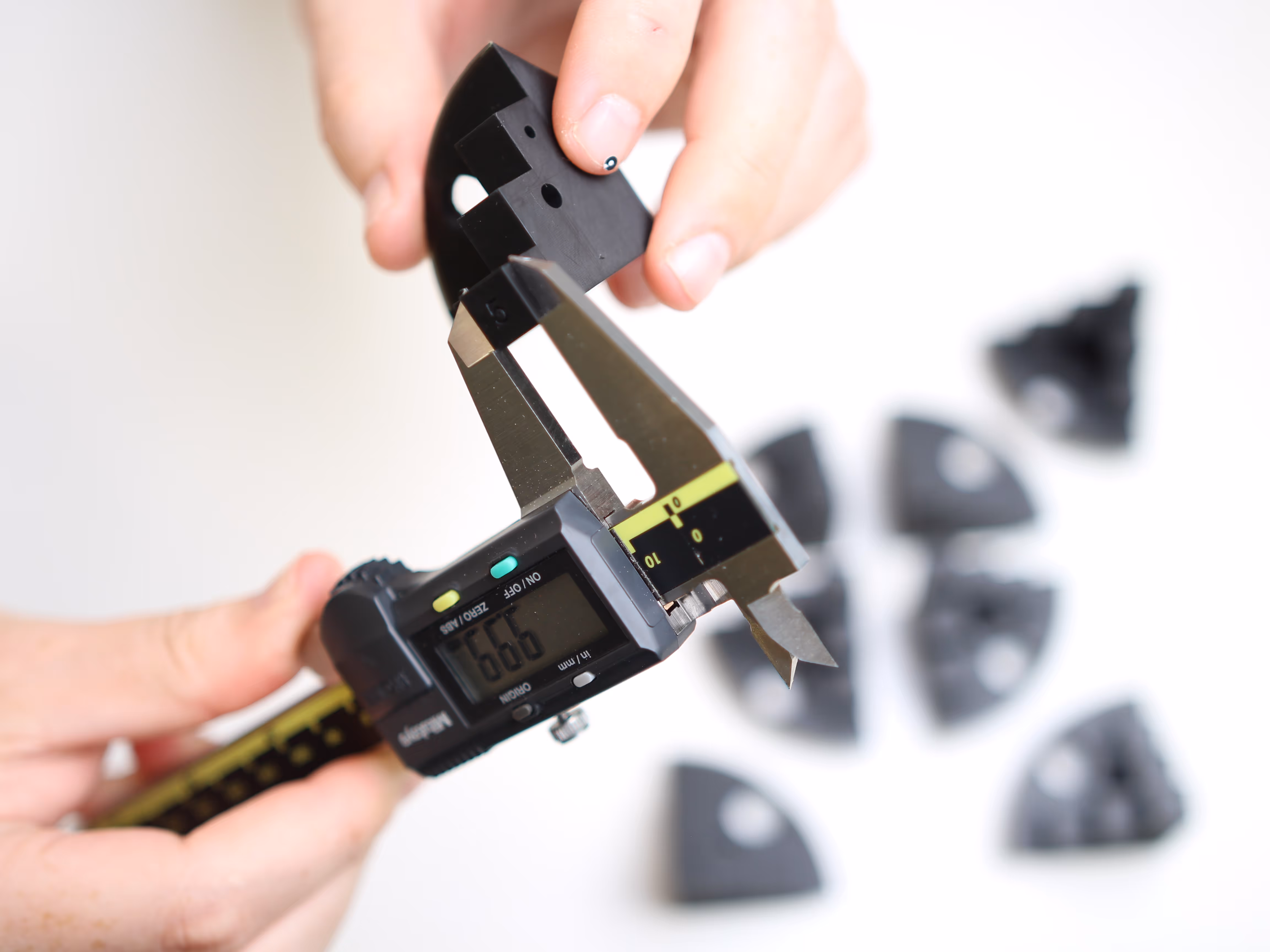 Hands holding a digital caliper measuring a small black plastic part with other similar parts blurred in the background.