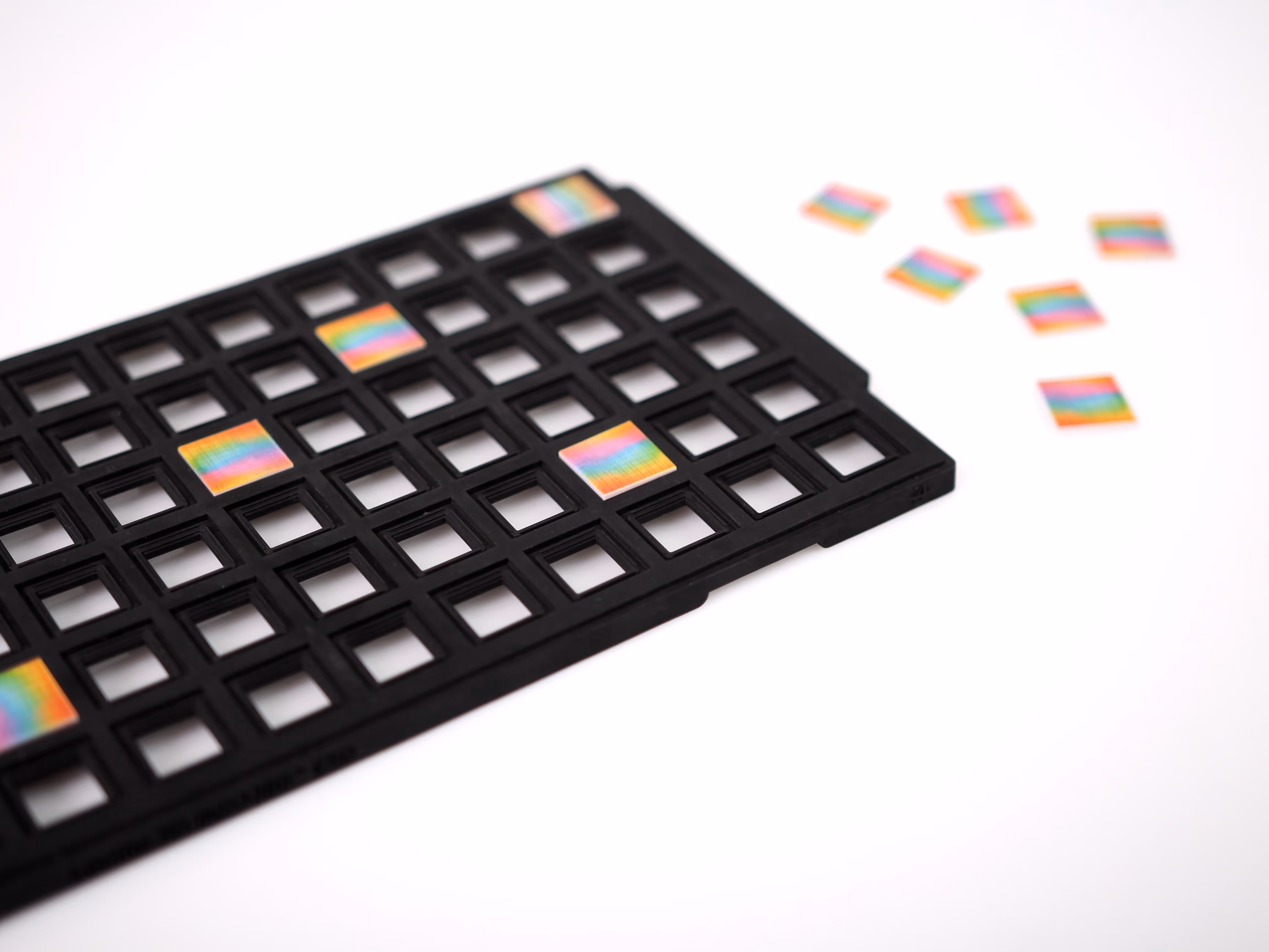 Black grid tray holding several small square chips with rainbow-colored reflective surfaces, with additional chips scattered beside it on a white surface.