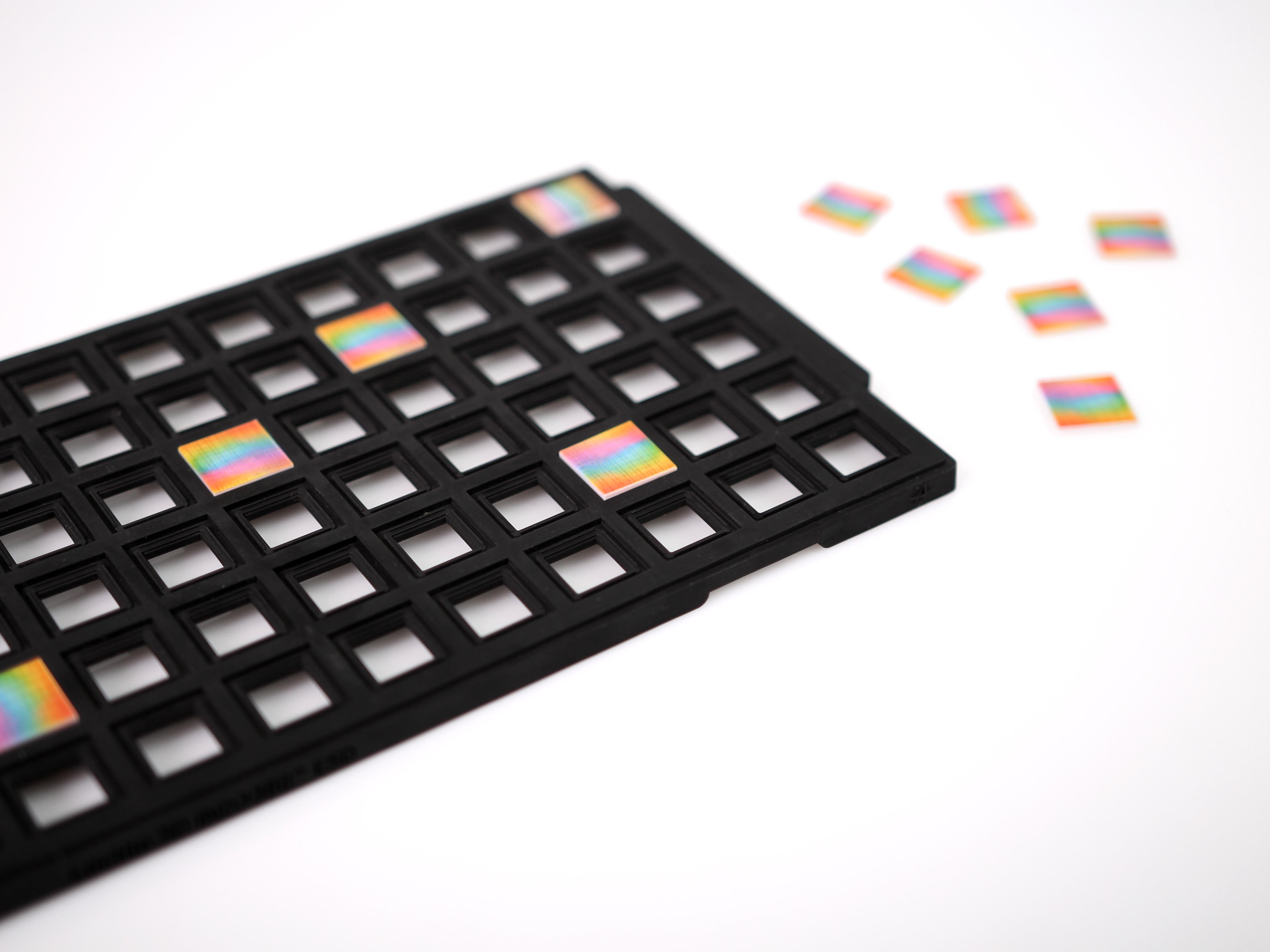 Black grid tray holding several small square chips with rainbow-colored reflective surfaces, with additional chips scattered beside it on a white surface.