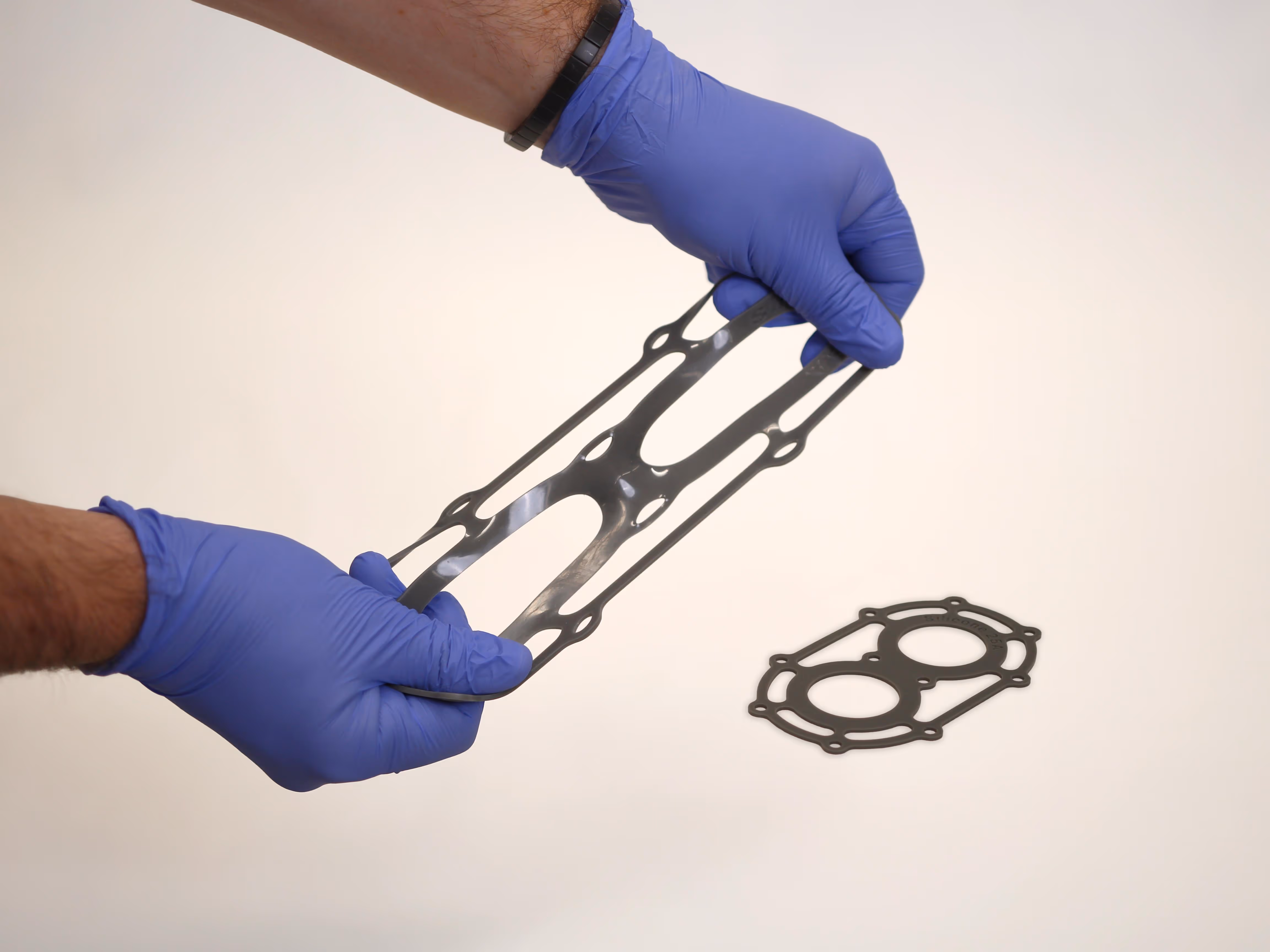 Person wearing blue gloves stretching a black gasket with another gasket placed on a white surface.