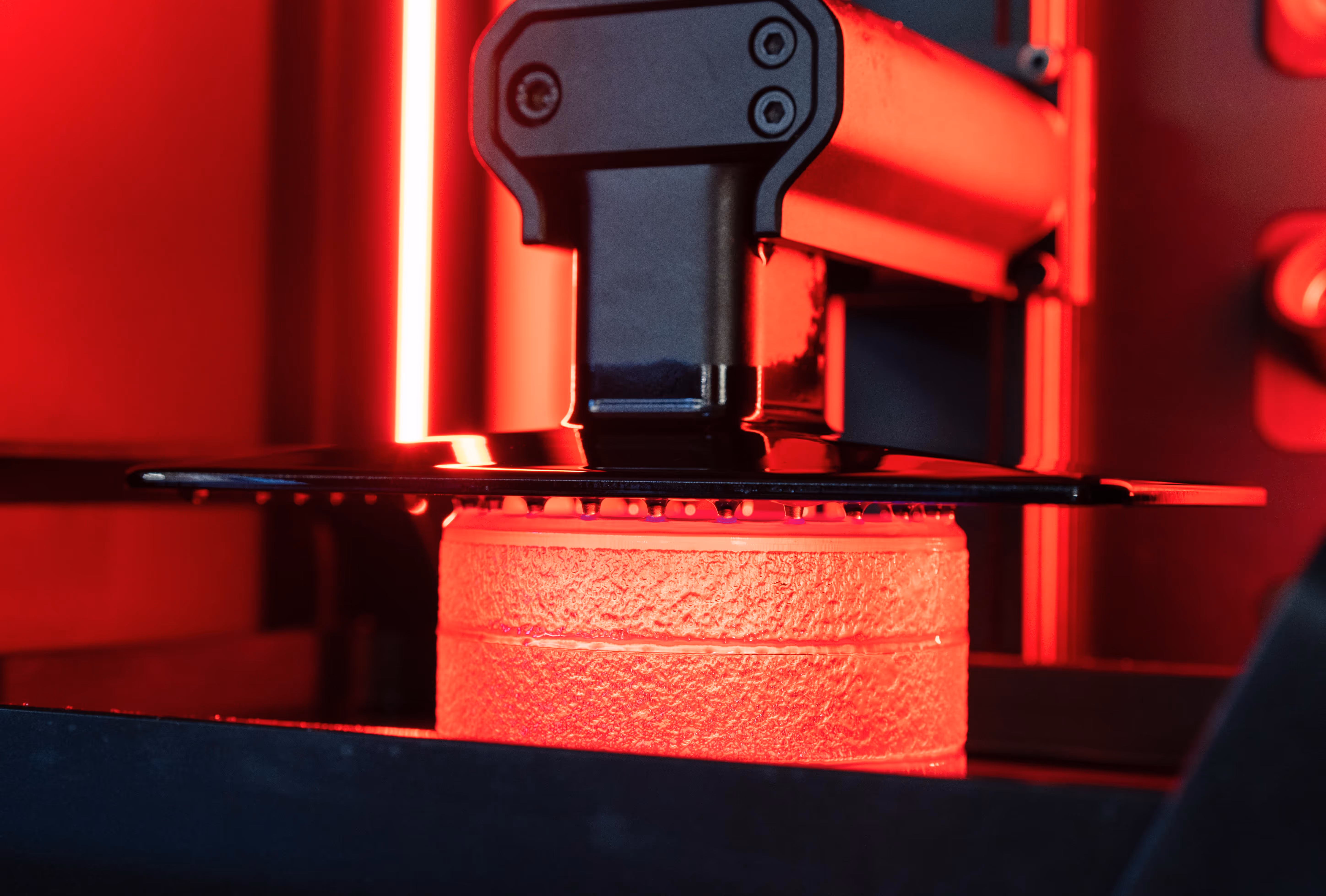 Close-up of a 3D printer creating a textured cylindrical object illuminated by red light.