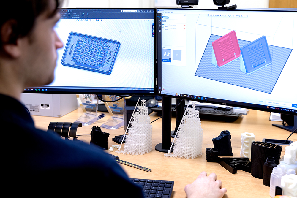 Person working at a desk with dual monitors displaying 3D modeling software and various 3D printed mechanical parts on the desk.