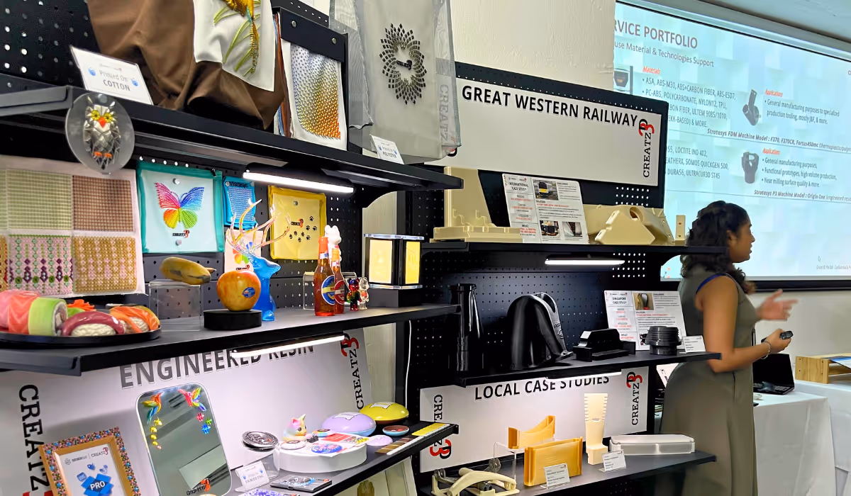 Woman giving a presentation next to shelves displaying colorful craft items, prototypes, and industrial design samples under signs like Great Western Railway, Engineered Resin, and Local Case Studies.
