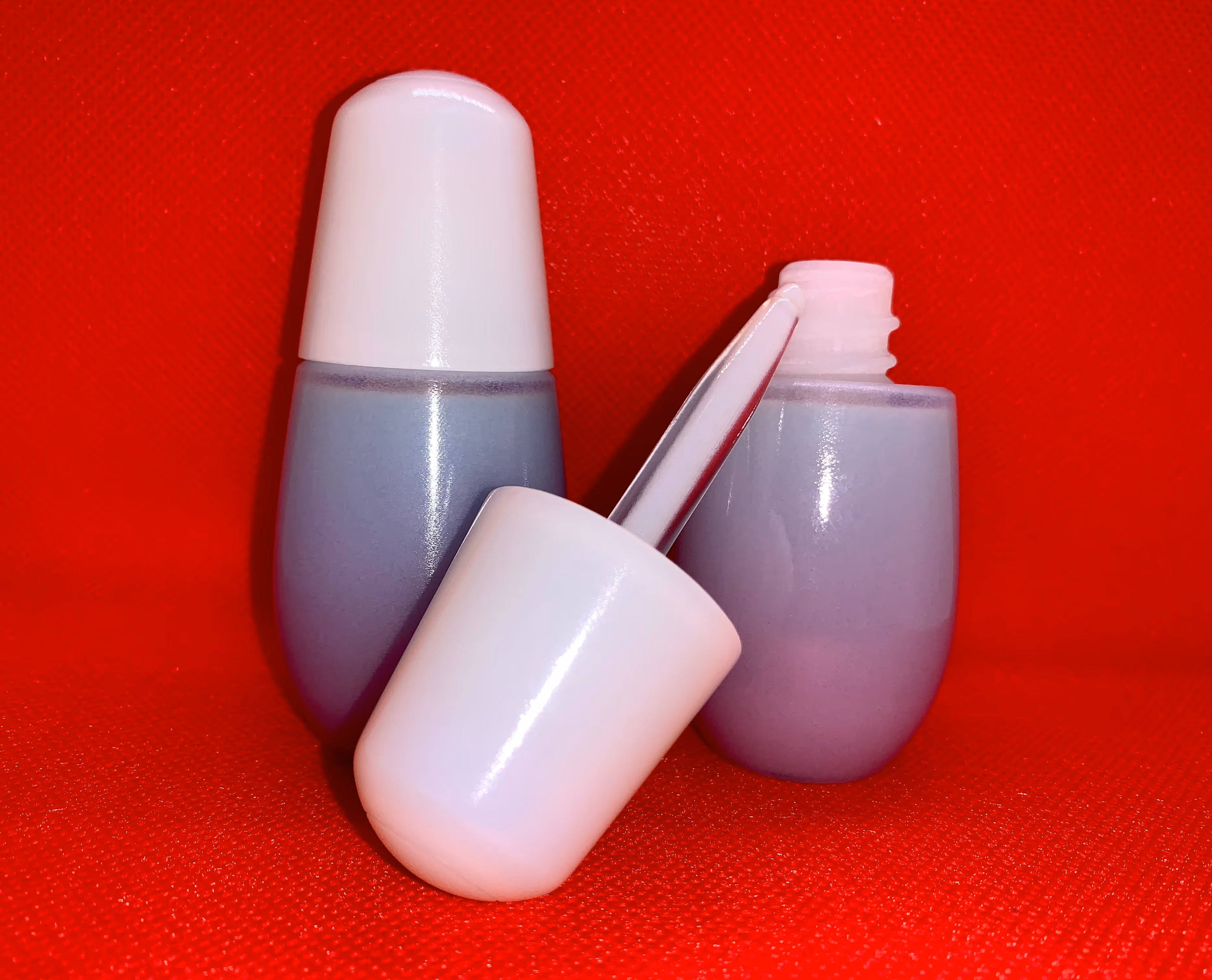Two small bottles of purple nail polish with white caps on a red textured surface, one bottle open with the brush resting beside it.