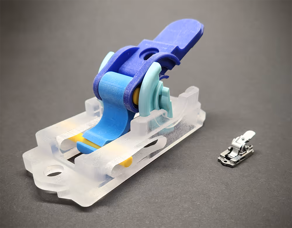 Close-up of a blue and transparent plastic hinge mechanism next to a small silver metal hinge piece on a gray surface.