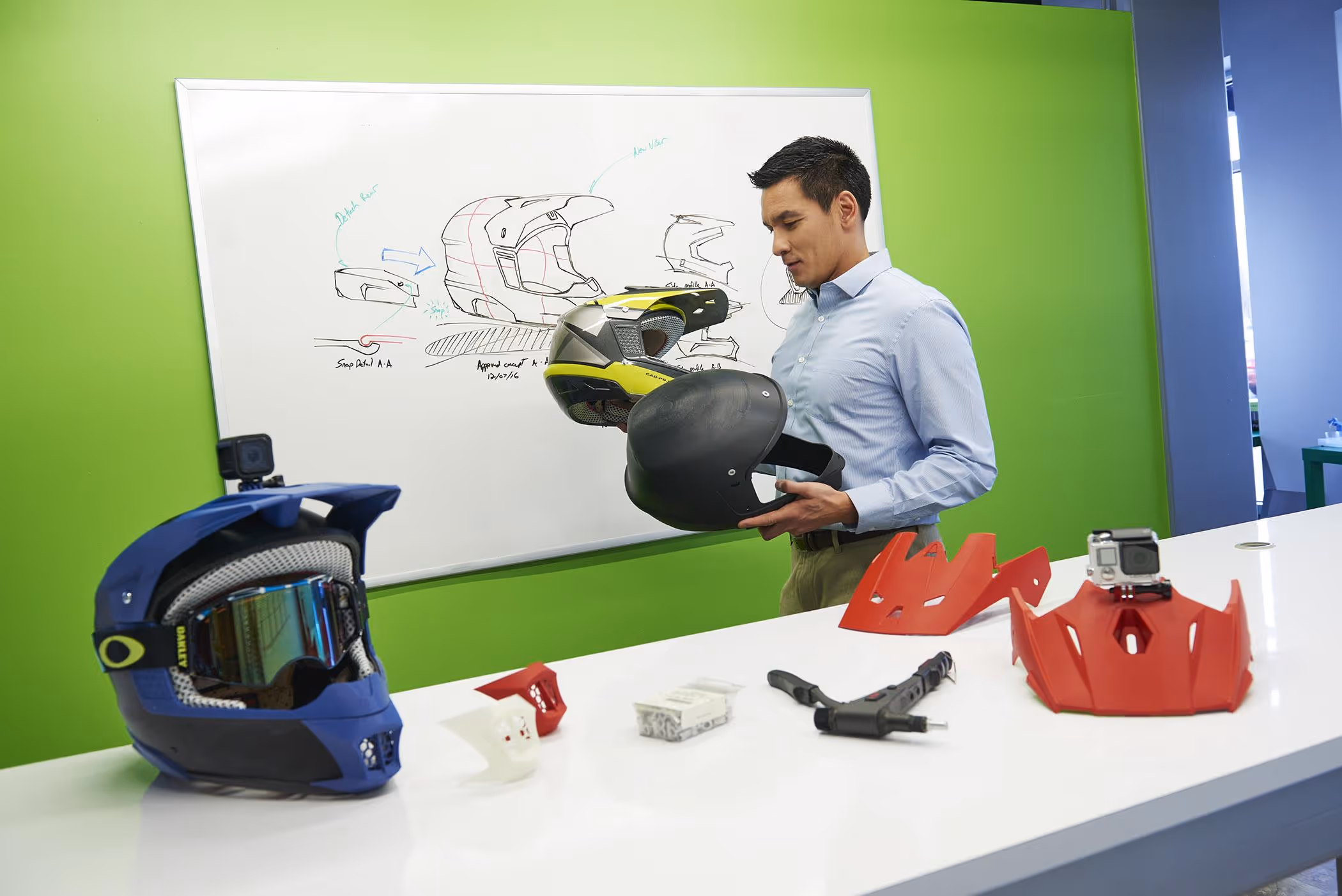 Man examining motorcycle helmet prototypes in an office with design sketches on a whiteboard and helmet parts on a table.
