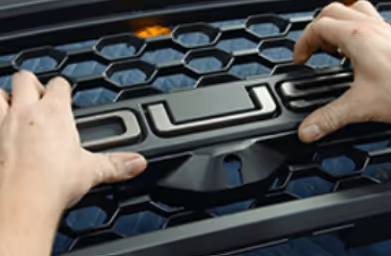 Person installing or adjusting a black grille with a honeycomb pattern on a vehicle.