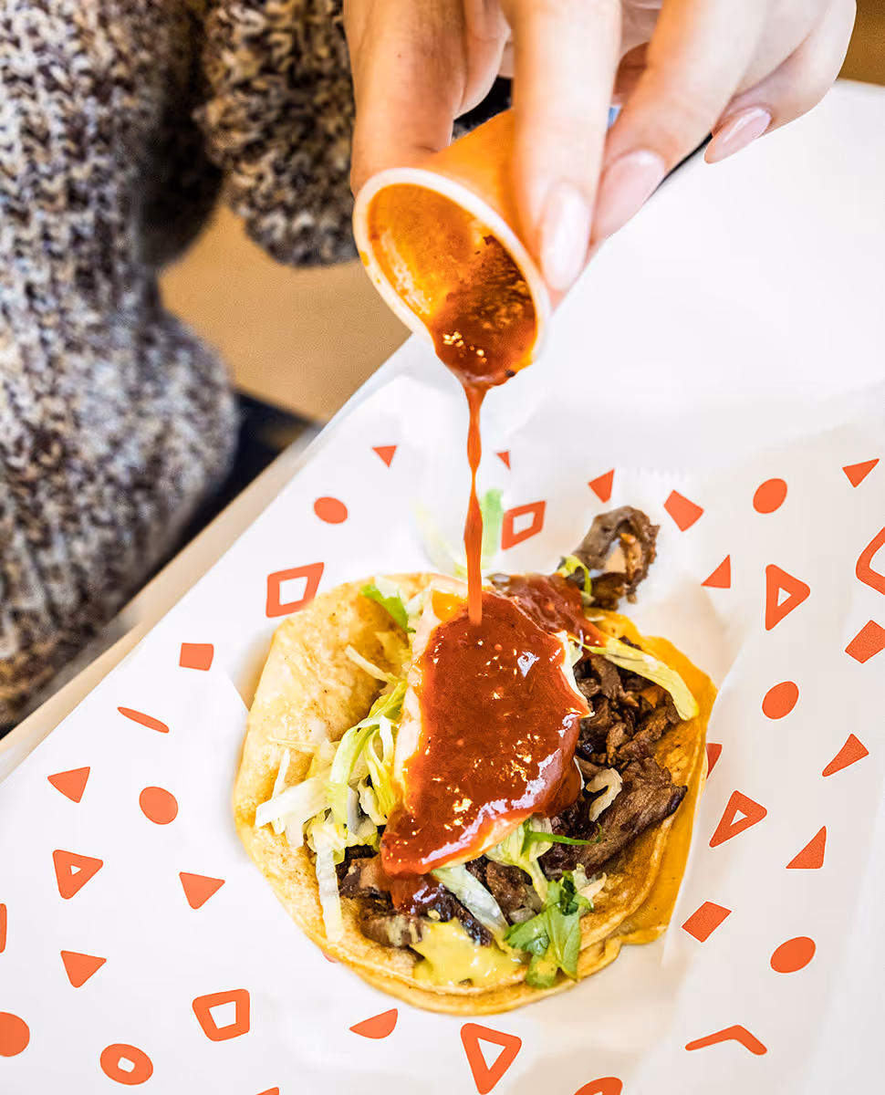 Person pouring spicy red salsa over a Mexican street taco with grilled beef, lettuce, and cheese at Tacos Yoyo, a taquería in Miami.