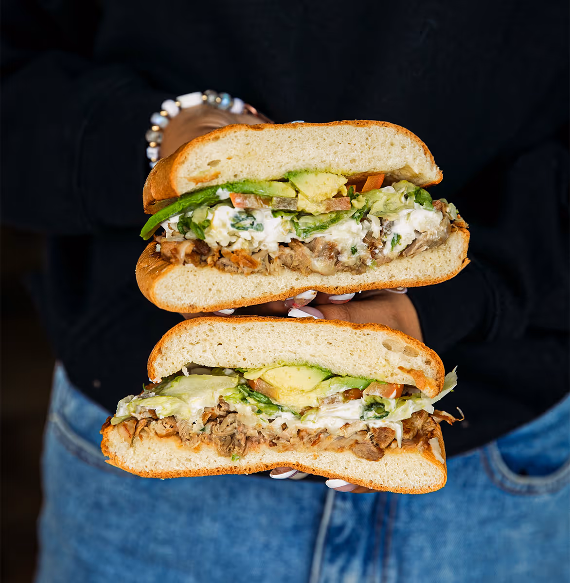 Person holding a Mexican torta sandwich cut in half, filled with shredded meat, avocado, lettuce, tomato, and crema, from Tacos Yoyo in Miami.