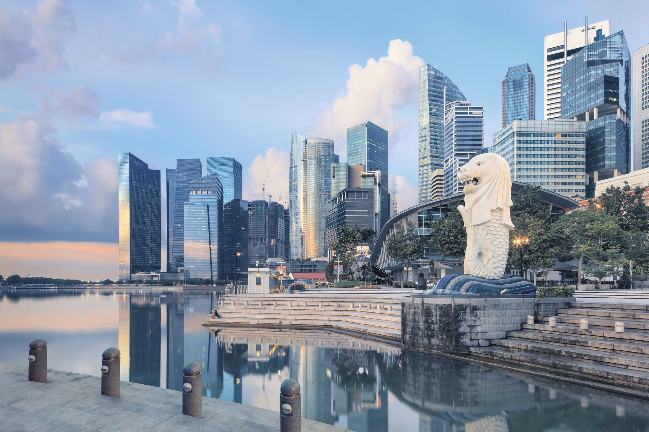 Panoramic view of Singapore’s financial district at sunrise, with the iconic Merlion statue in the foreground and modern skyscrapers reflected in the bay.