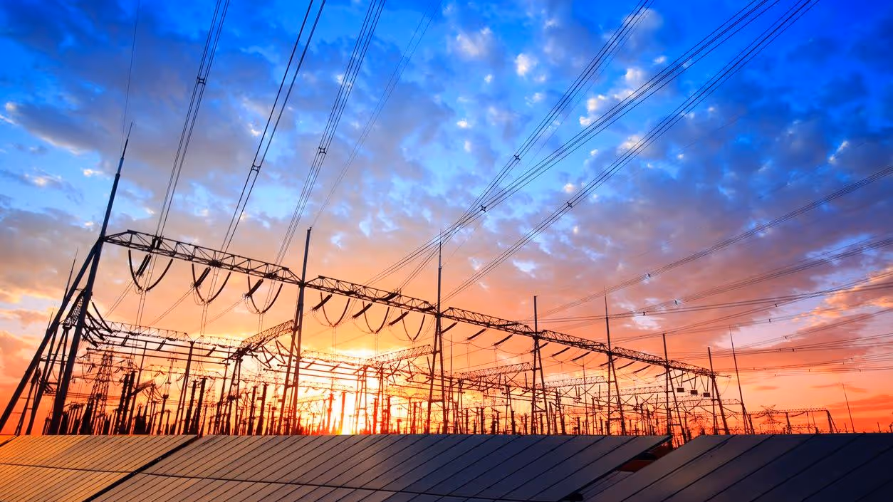 Electric power substation with transmission wires and solar panels under a colorful sunset sky.