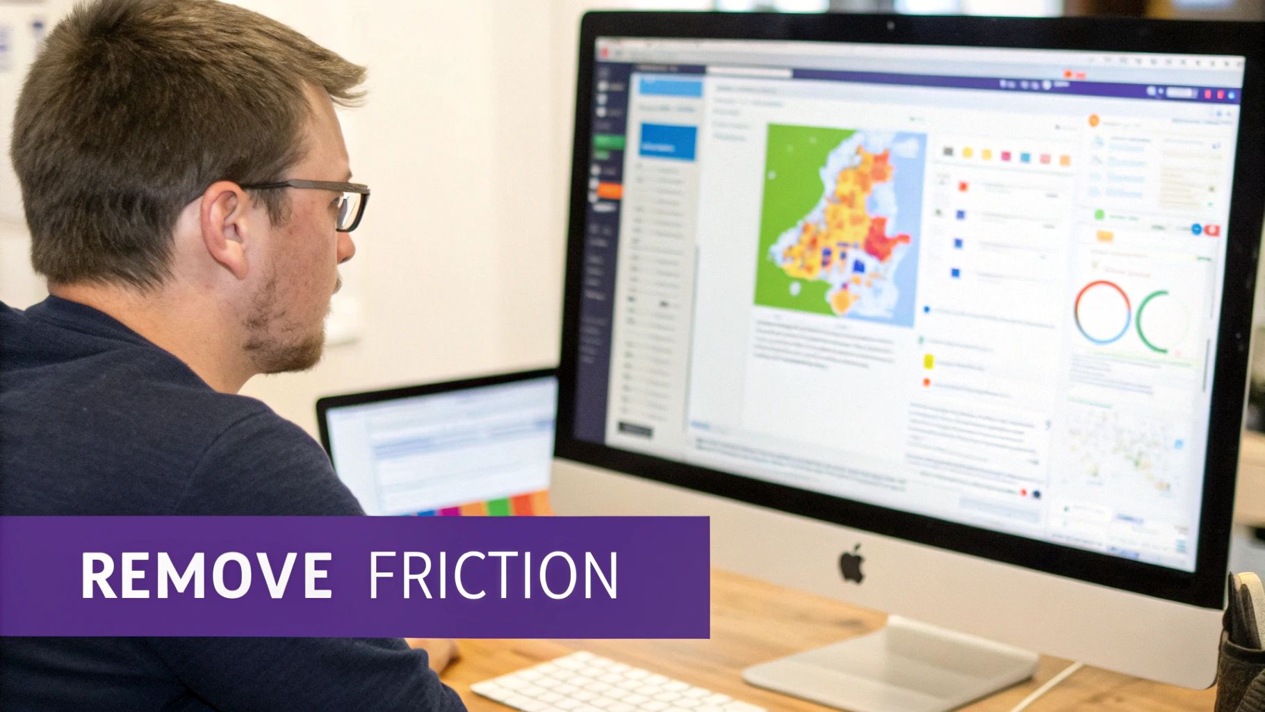 A man in glasses works on a desktop computer displaying data visualizations and charts.
