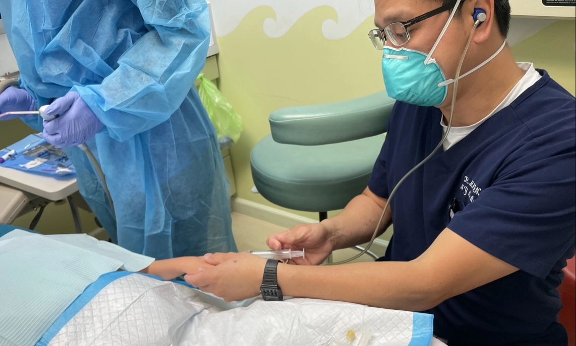 Pediatric dentist in protective gear administers sedation through an IV to a child patient during a dental procedure.