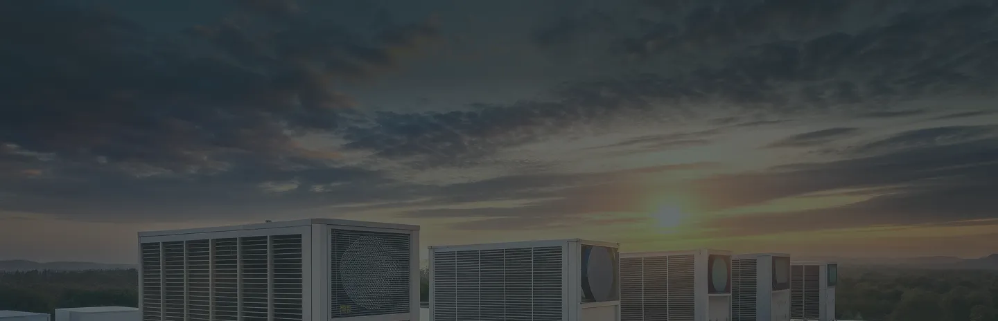 A row of large air conditioning units on a rooftop at sunset with a cloudy sky and distant tree-covered hills in the background.