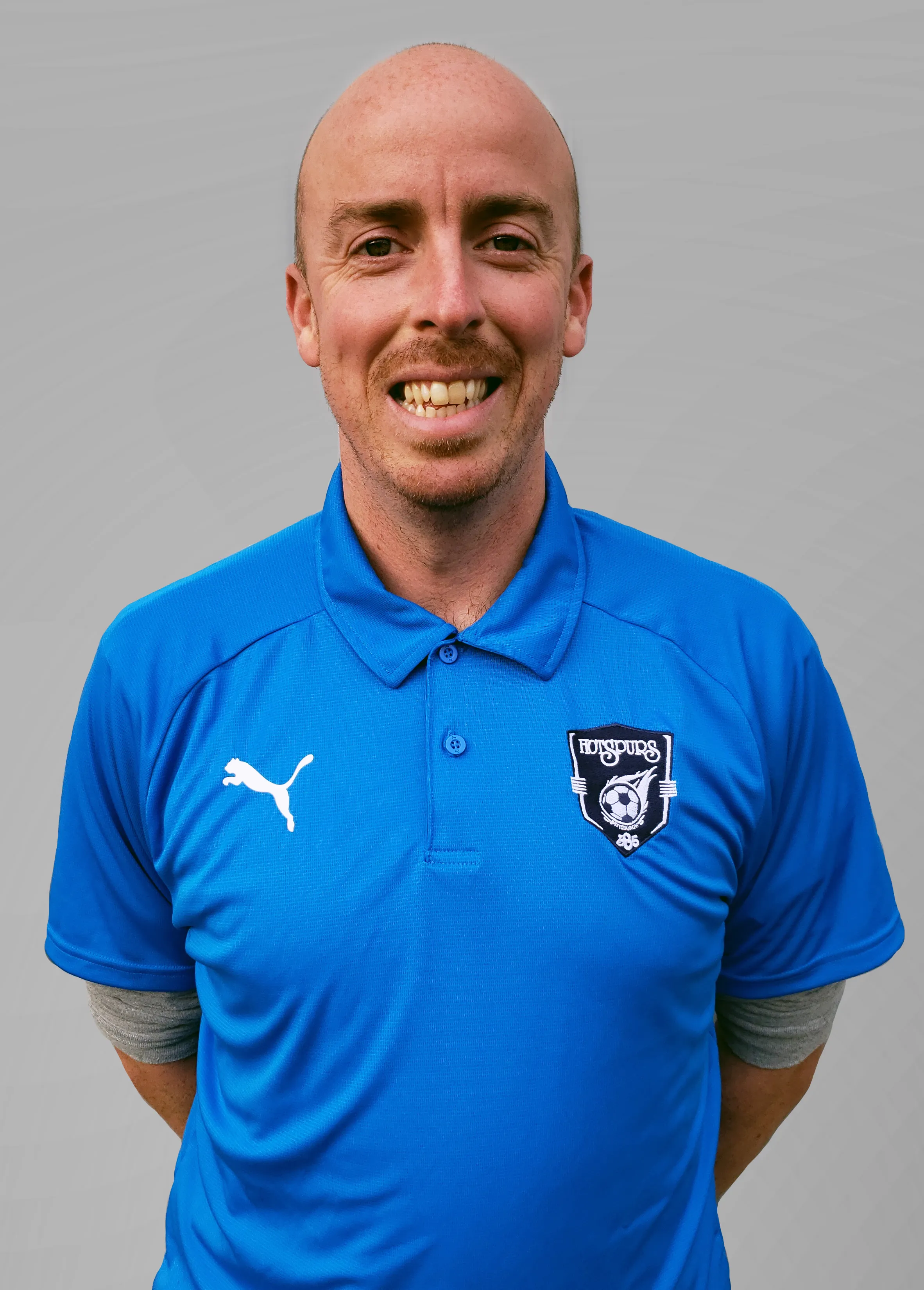 Smiling bald man wearing a blue Puma polo shirt with Hotspurs SC logo on the chest against a plain gray background.