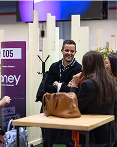 A man and three women engaged in conversation near a conference booth, with a bag on the table.