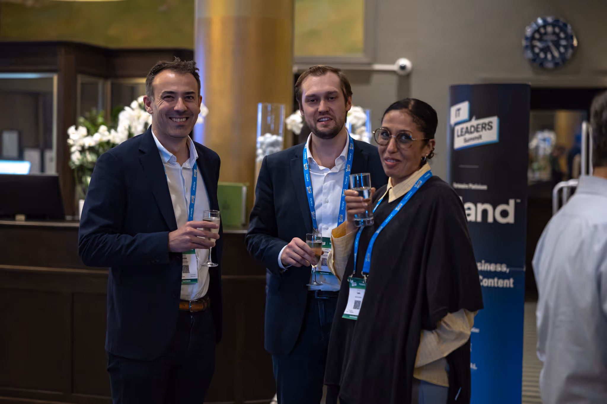 Three people standing together, smiling and holding drinks at a networking event indoors.