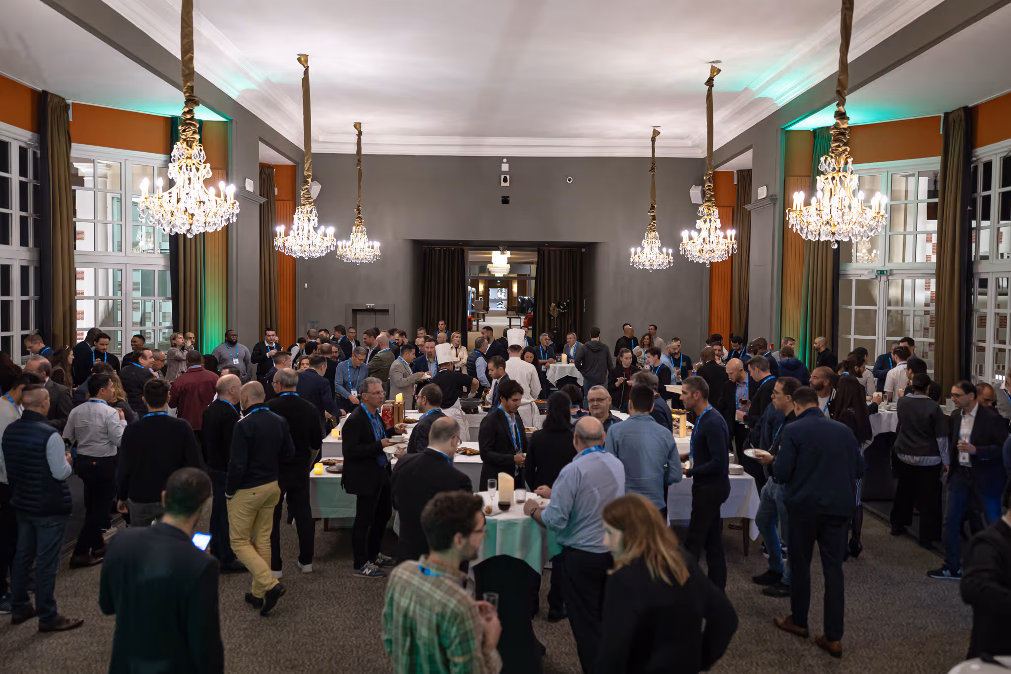 A large group of people mingles at a formal indoor event with chandeliers overhead.