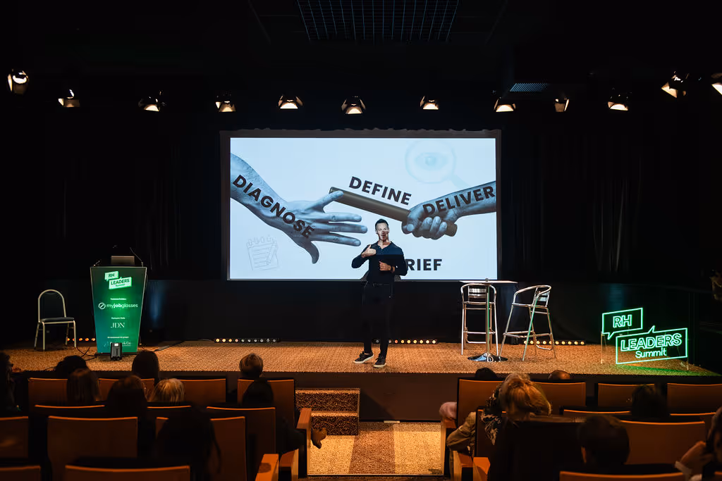 Man on stage giving a presentation with a large screen displaying words: Diagnose, Define, Deliver.