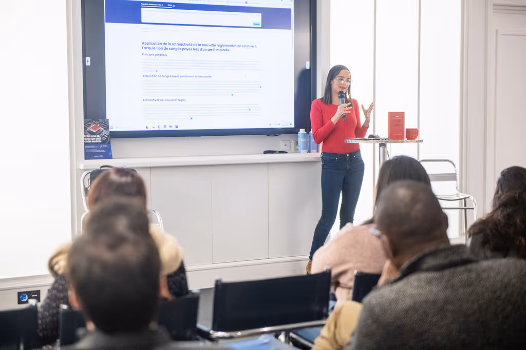 A woman in a red sweater presents to a seated audience using a large screen.