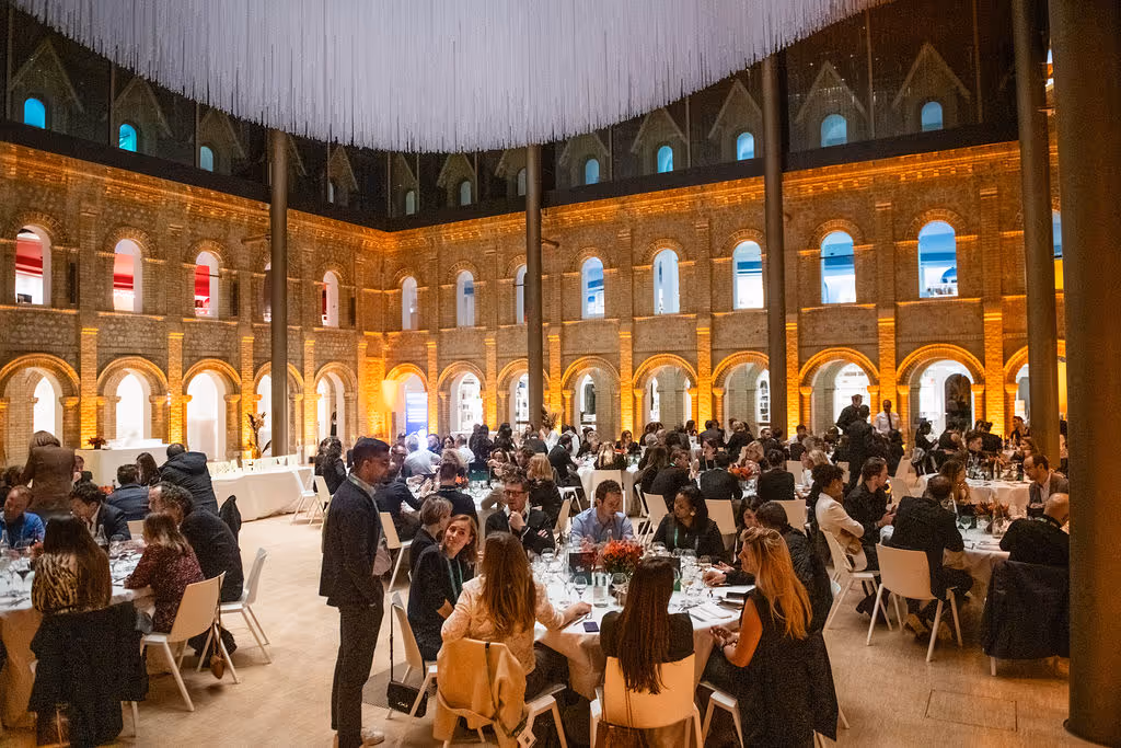 People seated at tables, dining and socializing, inside a large, illuminated hall with arches.