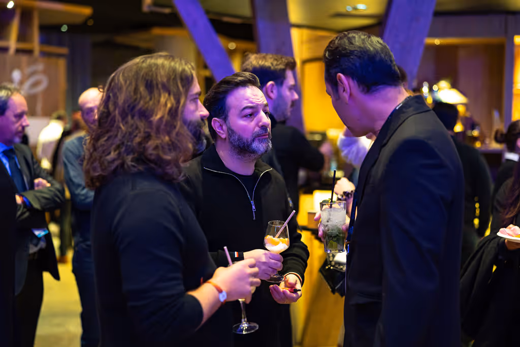 Three men stand talking at a social event, each holding a drink in their hand.